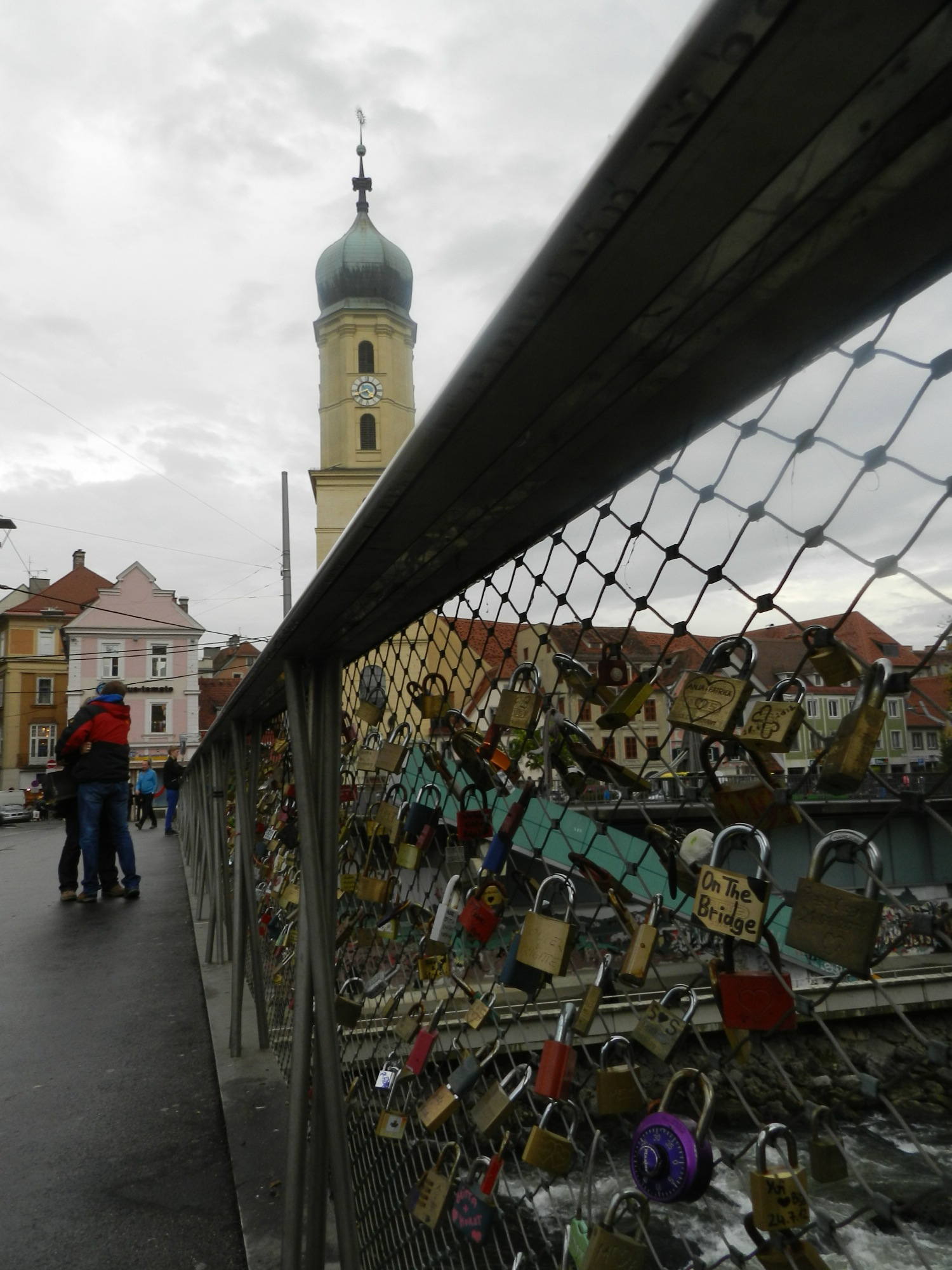Blick entlang des mit Schlössern geschmückten Geländers der Brücke mit Kirchtum und umarmenden Paar im Hintergrund