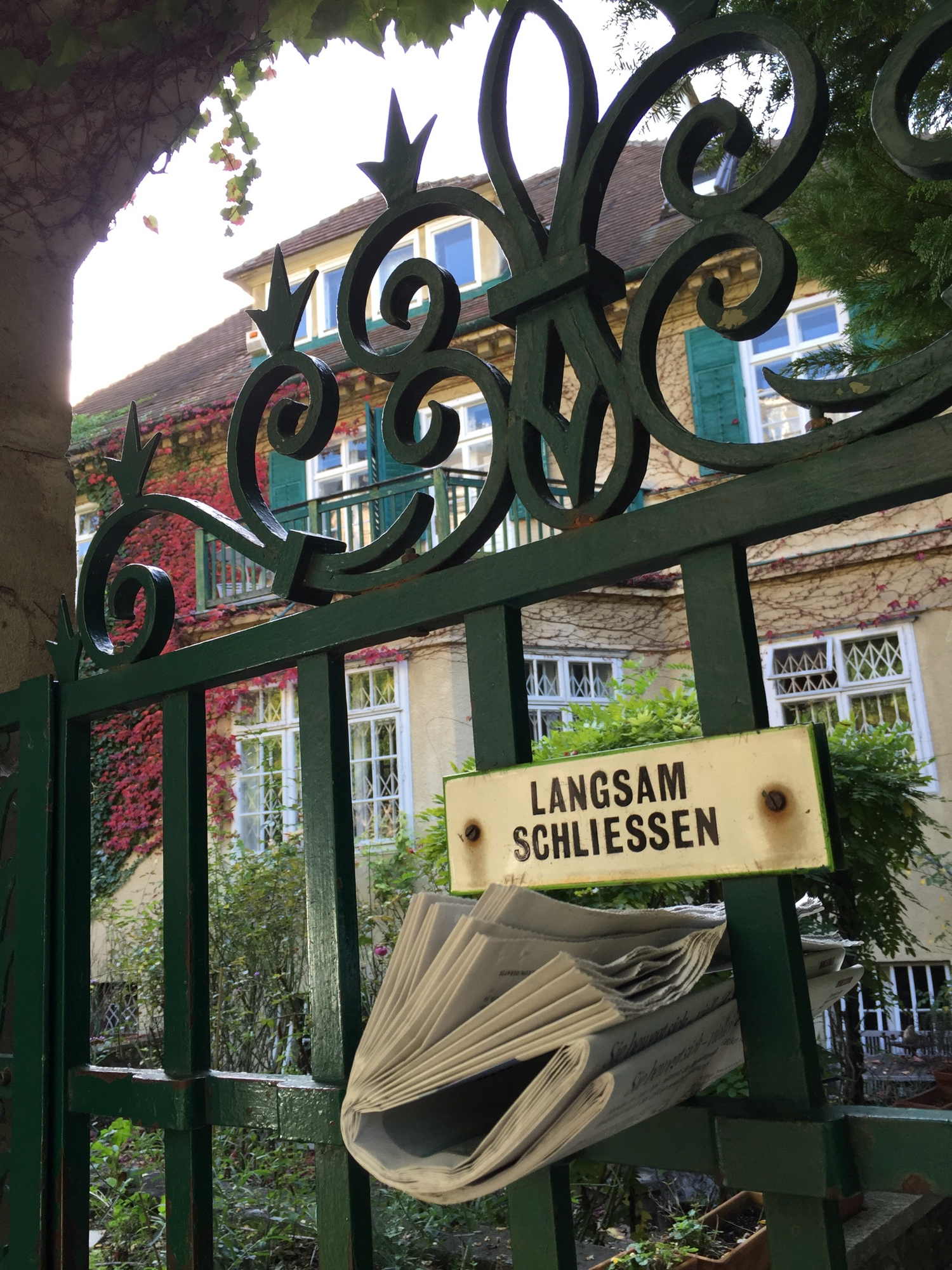 Gittertür vor Villa mit eingesteckter Zeitung und Schild „Langsam schließen”