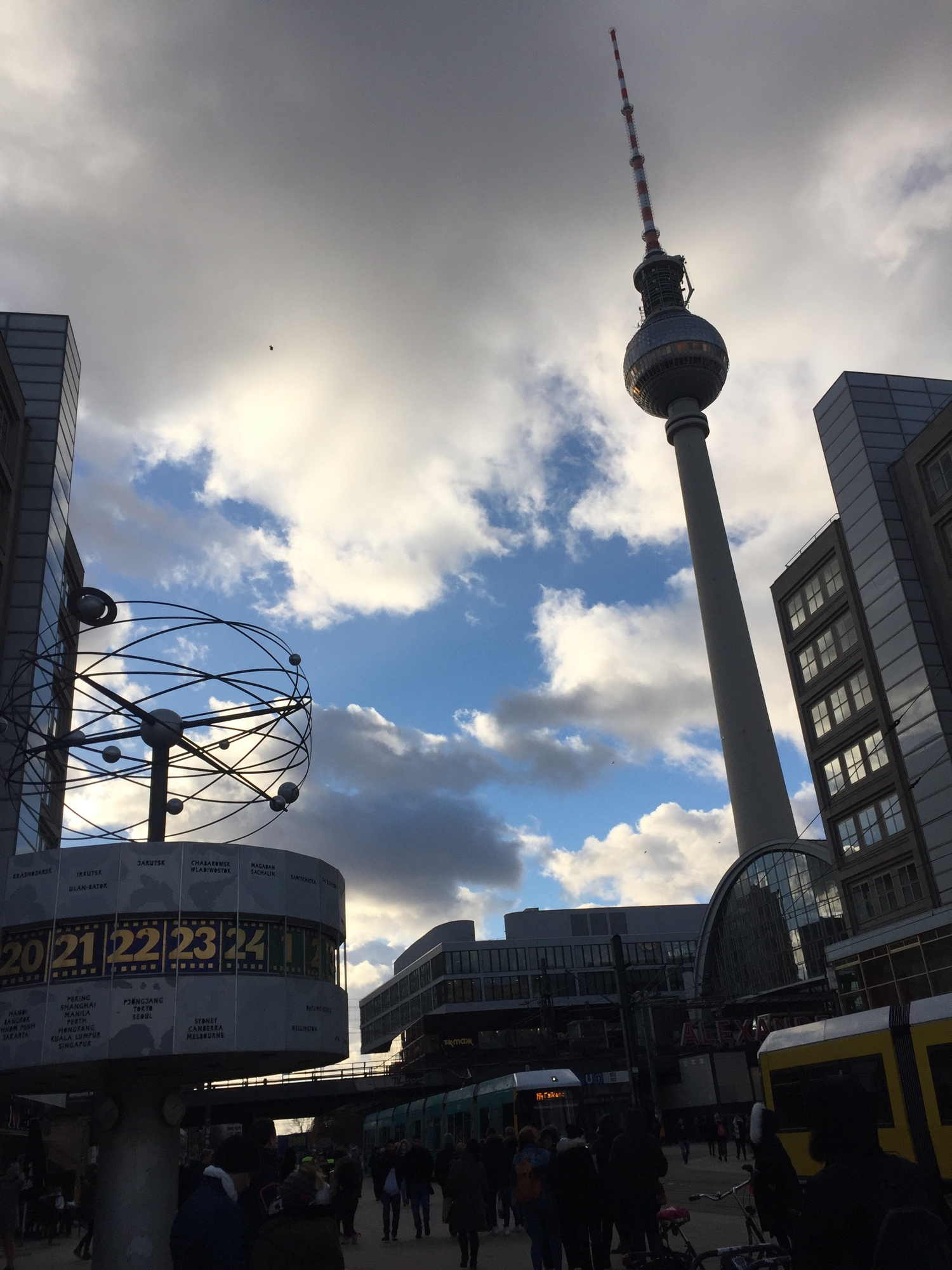 Alexanderplatz, Weltzeituhr links im Vordergrund, Fernsehturm im Hintergrund vor wolkigem Himmel