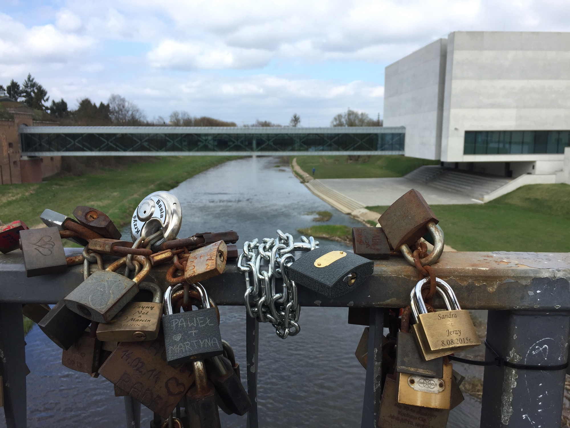 im Vordergrund Lovelocks/Liebesschlösser an einem Brückengeländer, im Hintergrund das moderne Gebäude des Kulturzentrums Brama Poznania