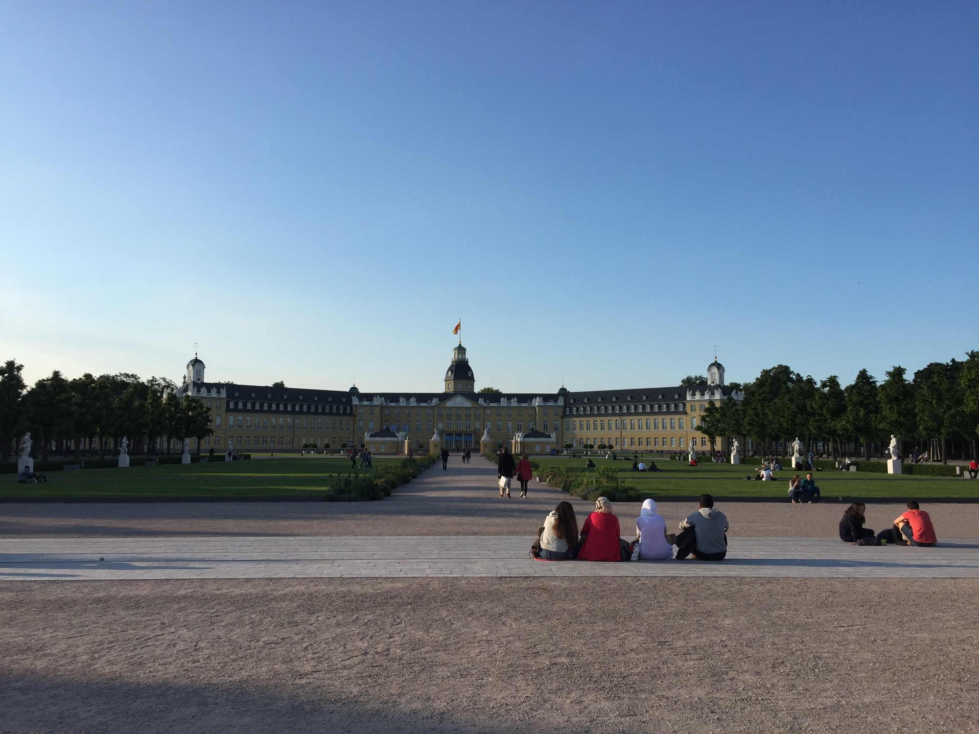 Blick auf das Schloss Karlsruhe, von dem ausgehend sich fächerförmig die Innenstadt entfaltet, im Vordergrund sitzen Jugendliche mit dem Rücken zur Kamera auf einer Treppe in der Sonne