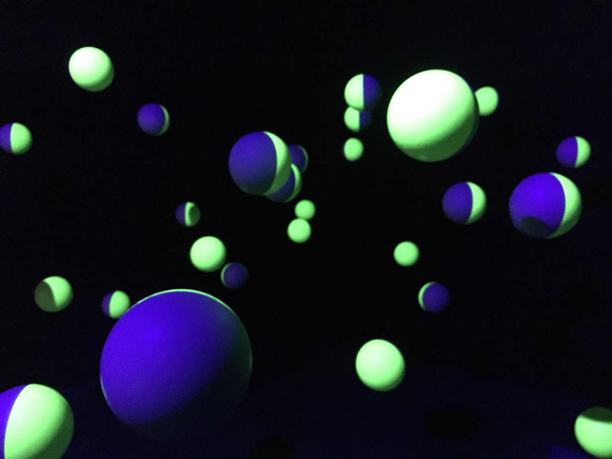 Installation mit von der Decke hängenden Styroporkugeln in einem dunklen Raum, die Kugeln sind je zur Hälfte mit verschiedenen UV-empfindlichen Farben bemalt