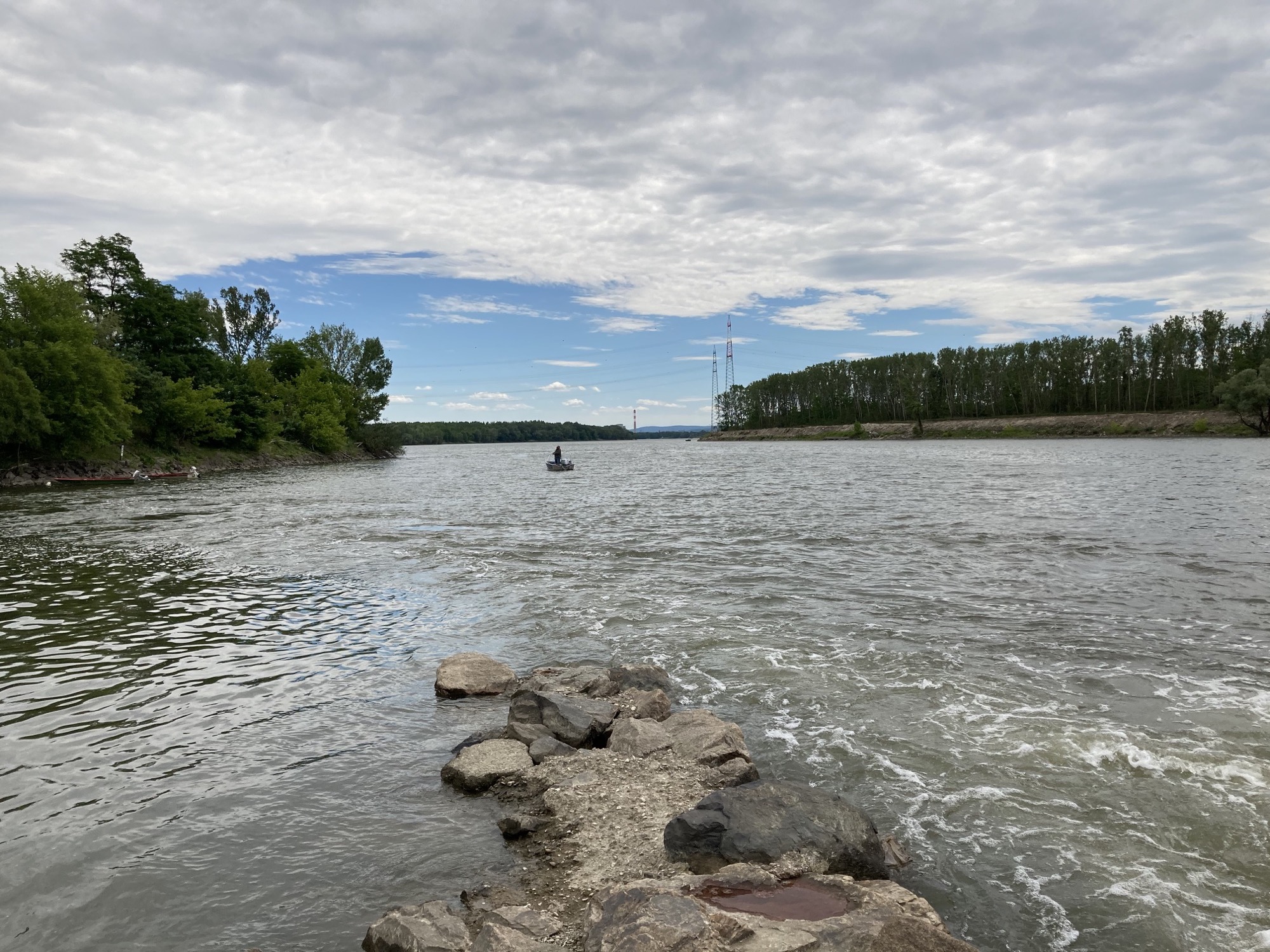 Mündung der Krems in die Donau, im Vordergrund eine ins Wasser ragende Steinspitze, dahinter ein Boot mit Angler, im Hintergrund Strommasten und der Turm eines Kraftwerks