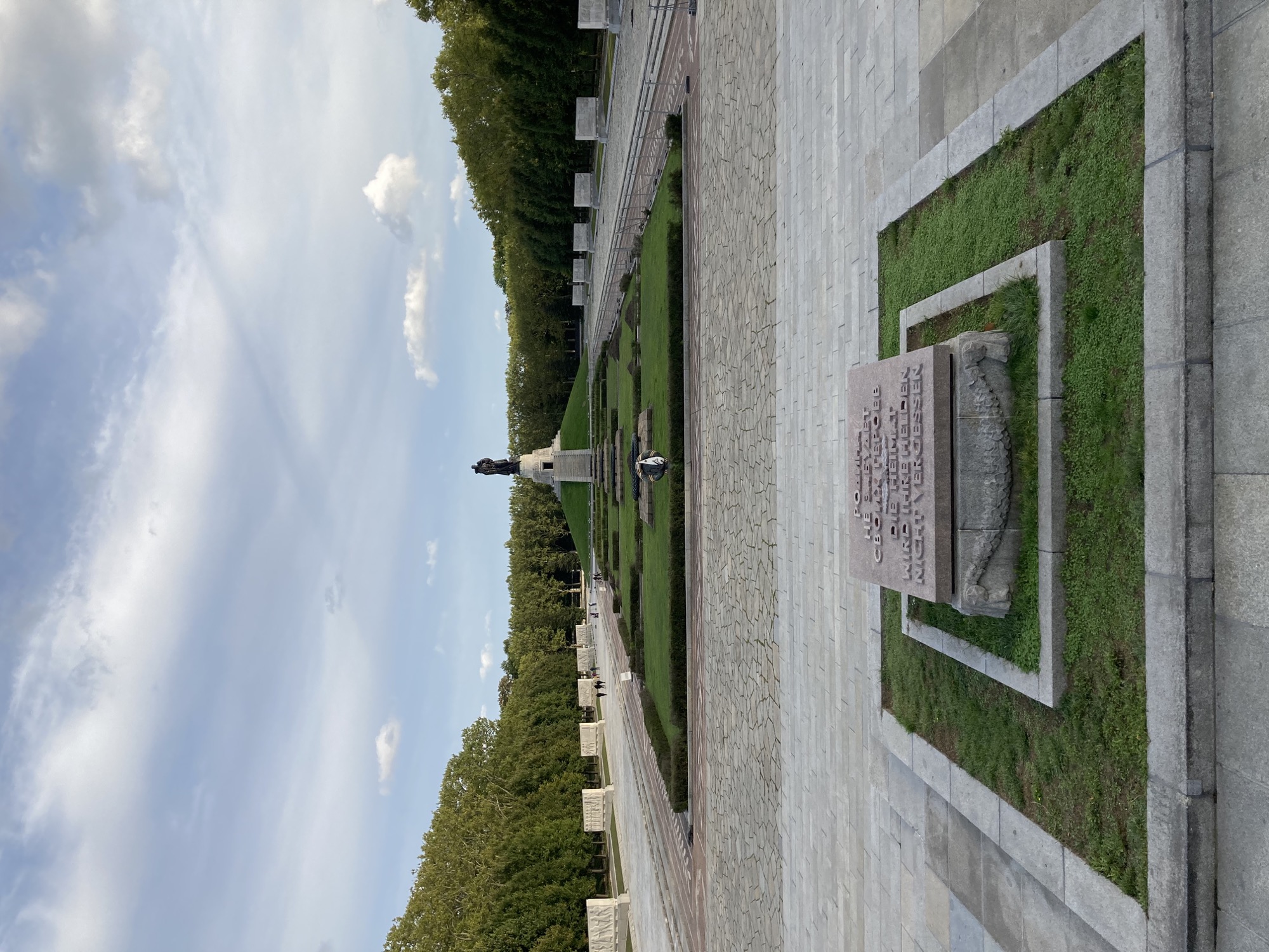 Weitblick über die Gedenkstätte des Sowjetischen Ehrenmals, im Vordergrund ein Gedenkstein mit der Inschrift „Die Heimat wird ihre Helden nicht vergessen“, im Mittelgrund ein Kranz mit Leuchtturm und Anker, im Hintergrund die große Statue des Soldaten mit Kind auf dem Arm