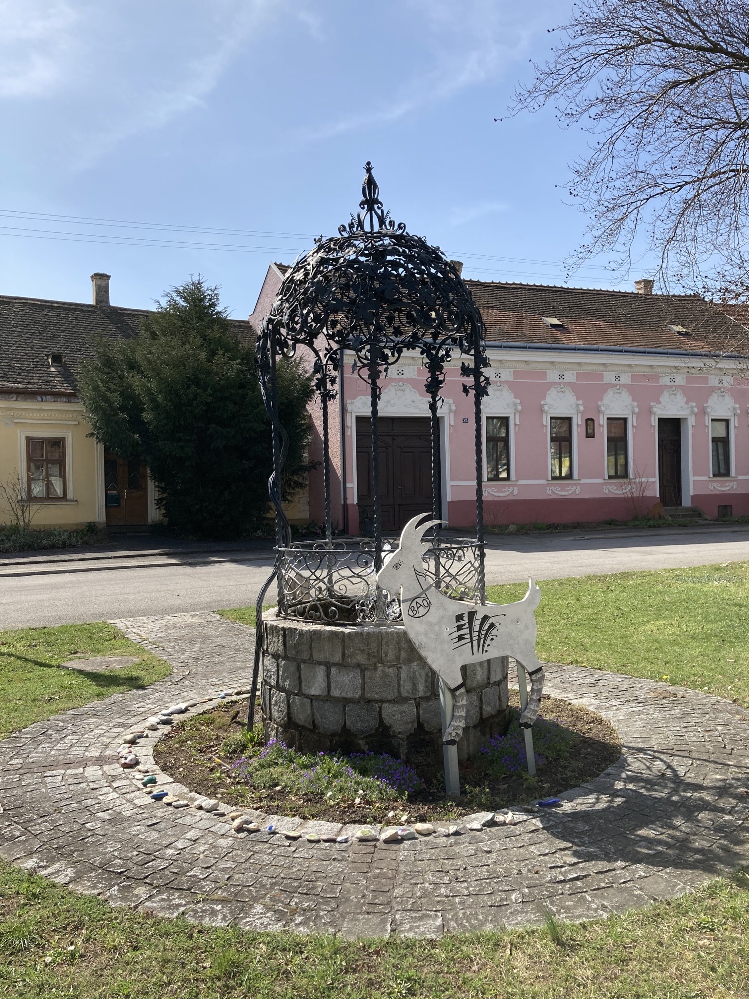 Brunnen im Zentrum von Bockfließ, mit dekorativem Blattdach aus Metall und davor einer Darstellung eines Bocks