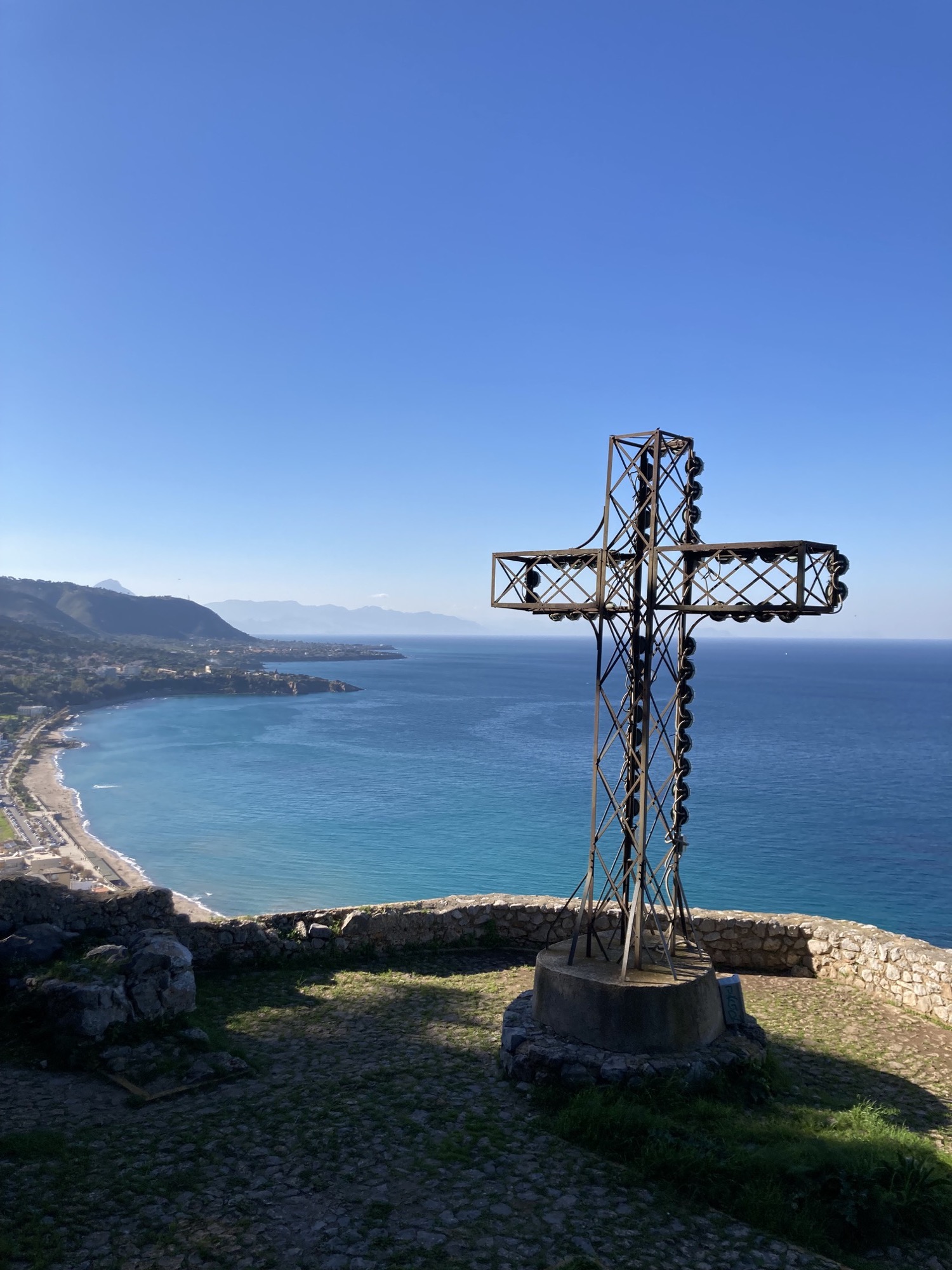 im Vordergrund ein großes christliches Kreuz, im Hintergrund unten der Strand von Cefalú