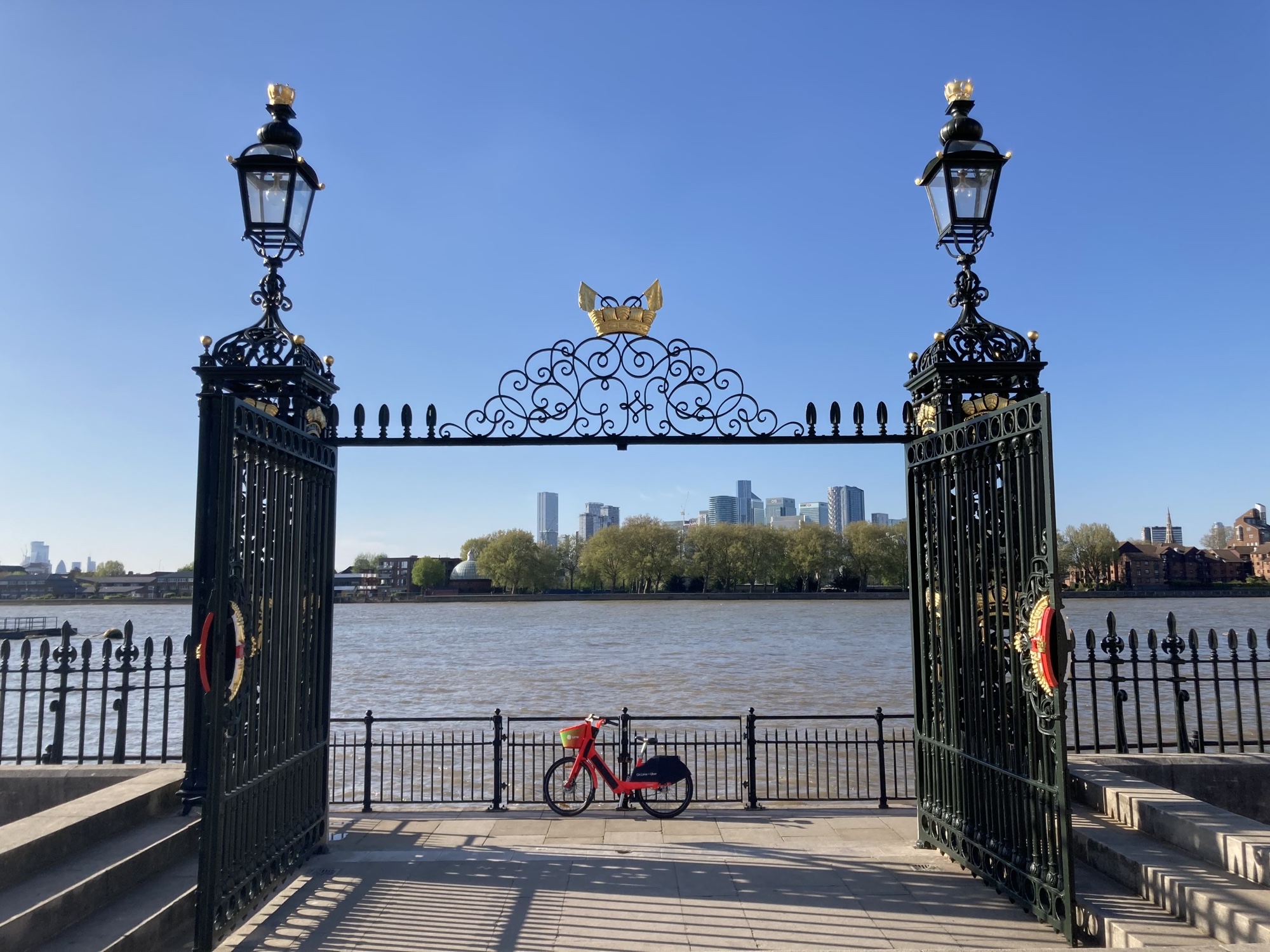 Blick auf die Themse und dahinter die Skyline von Zentrallondon durch ein geöffnetes schmiedeeisernes Tor, das links und rechts von imposanten Lampen und mittig von goldenen Schiffen und Flaggen geschmückt wird, auf dem Uferweg hinter dem Tor steht ein rotes Leihrad