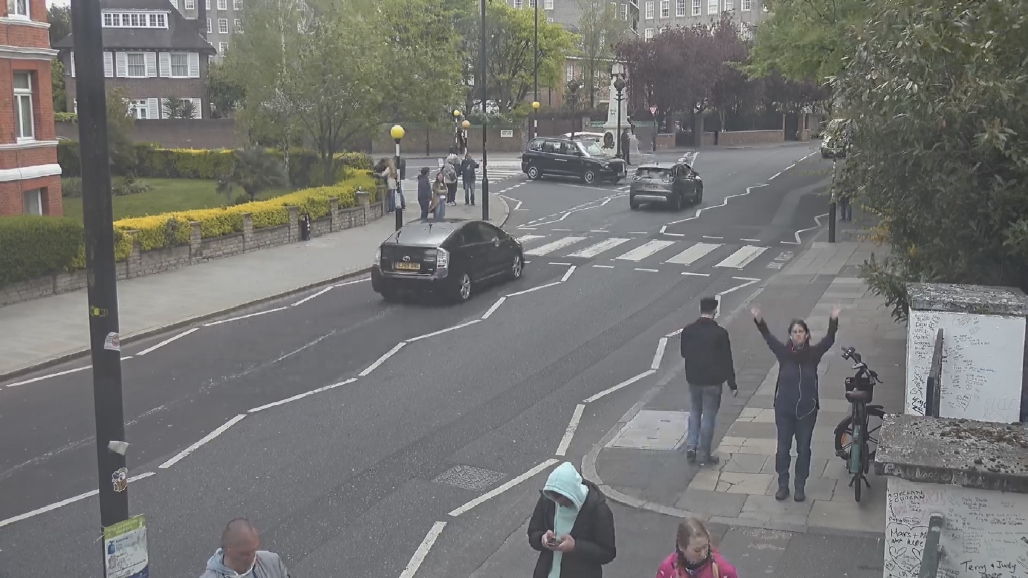 Webcam-Screenshot des berühmten Zebrastreifens vor den Abbey Road Studios, 4 Autos sind unterwegs, Menschen warten darauf, die Straße zu überqueren, im Hintergrund eine Statue, rechts vorne neben einem Fahrrad winkt eine Person (ich) in die Kamera