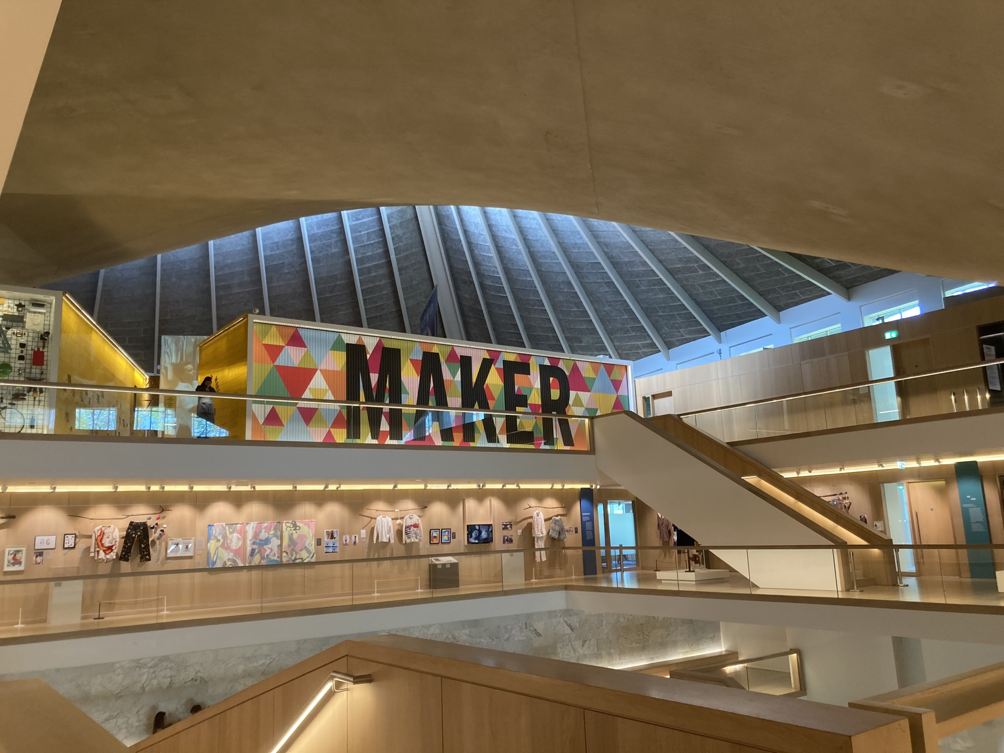 Blick durch den offenen Foyerraum des Design Museums, oben scheint Licht durch die Glaskuppel herein, im Mittelgrund eine Textwand mit dem Schriftzug „Maker“, von dort führt eine Treppe ein Stockwerk tiefer, wo Kleidung aus der Modeausstellung zu sehen ist
