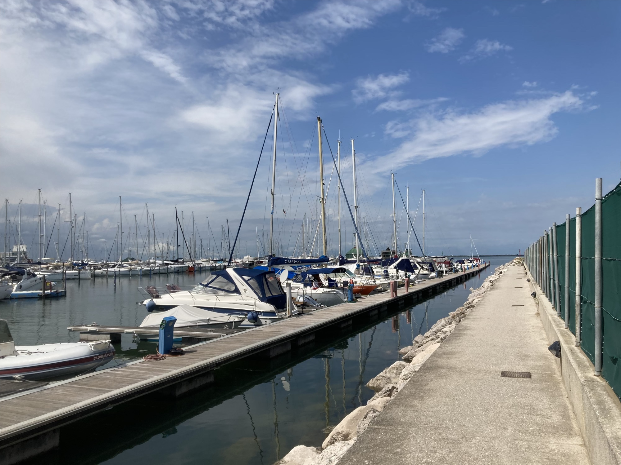 Hafen von Lignano, rechts ein geradeaus führender Weg am Wasser entlang, links verschiedene geparkte Boote und Segelboote
