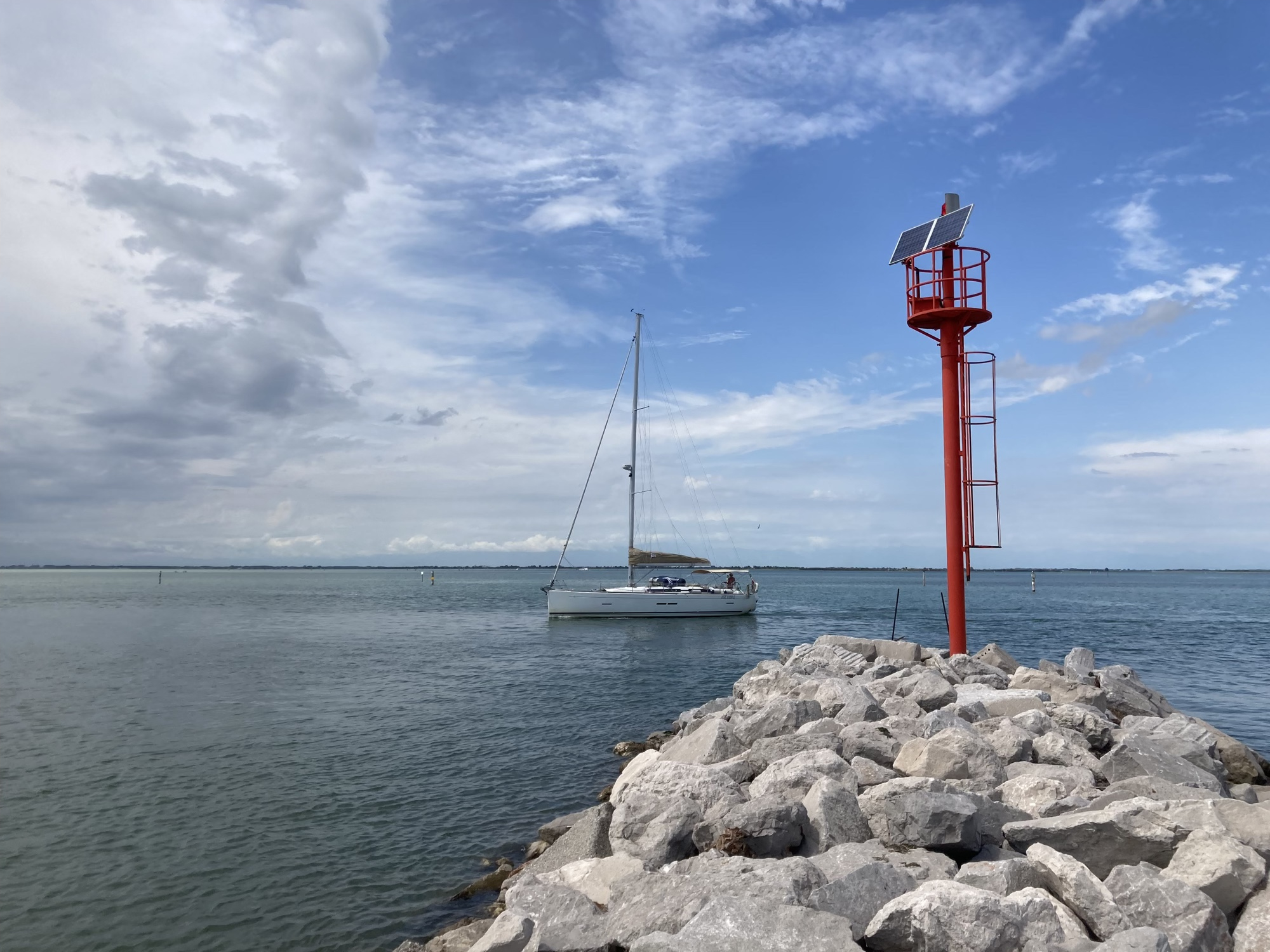 der nördlichste Punkt von Lignano, ein rot gestrichener Turm, gekrönt mit einer Solaranlage auf einer Landzunge aus Steinen, dahinter im Meer ein Seegelboot