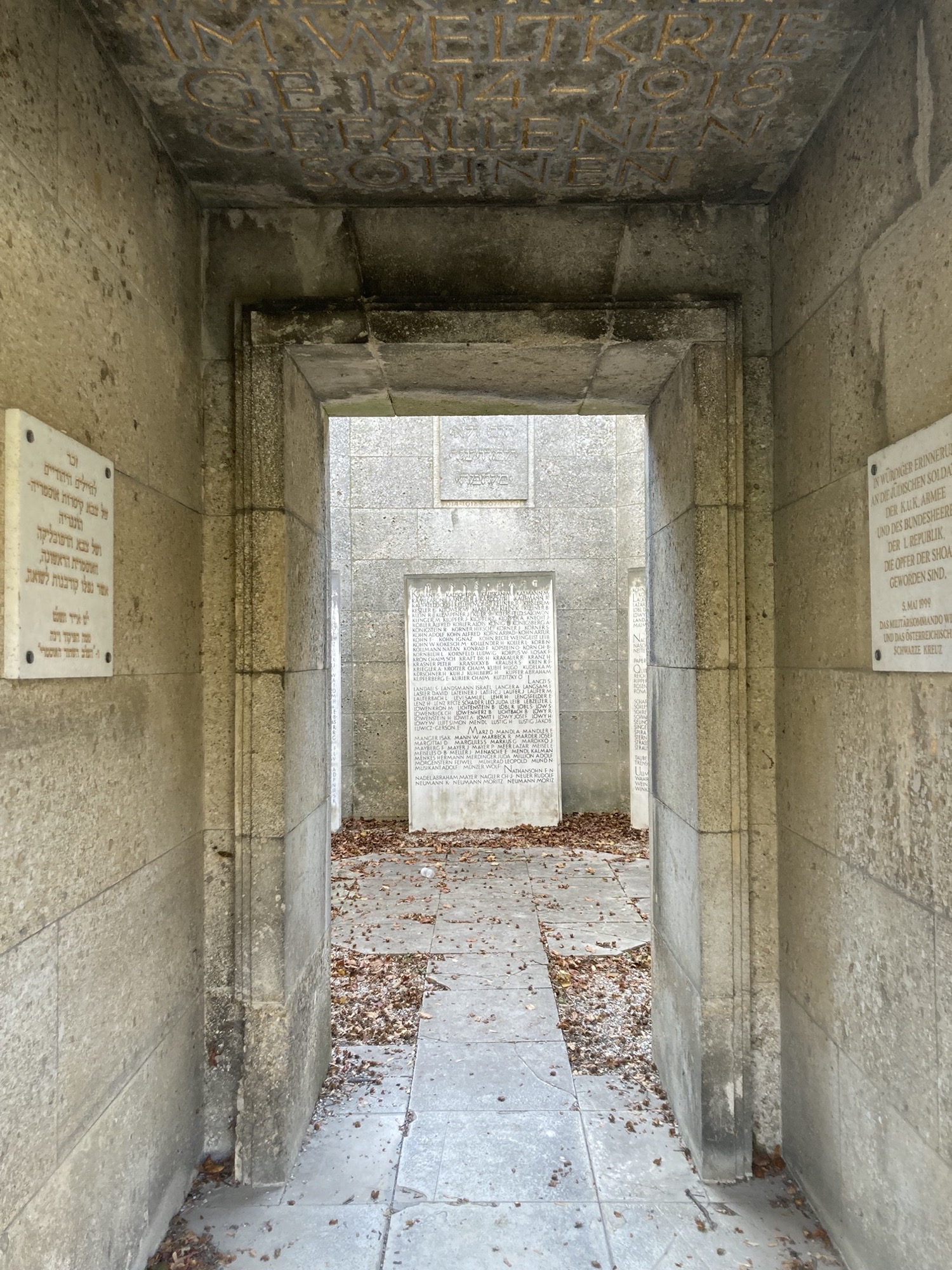 Blick durch den Zugang einer Gedenkstätte der Israelitischen Kultusgemeinde für die gefallenen des ersten Weltkriegs, links und rechts an der Wand weiße Steinfafeln, links mit hebräischen Schriftzeichen, rechts mit der deutschen Übersetzung, innerhalb der Gedenkstätte sind weitere große Steintafeln zu sehen, der Boden ist mit Herbstlaub bedeckt