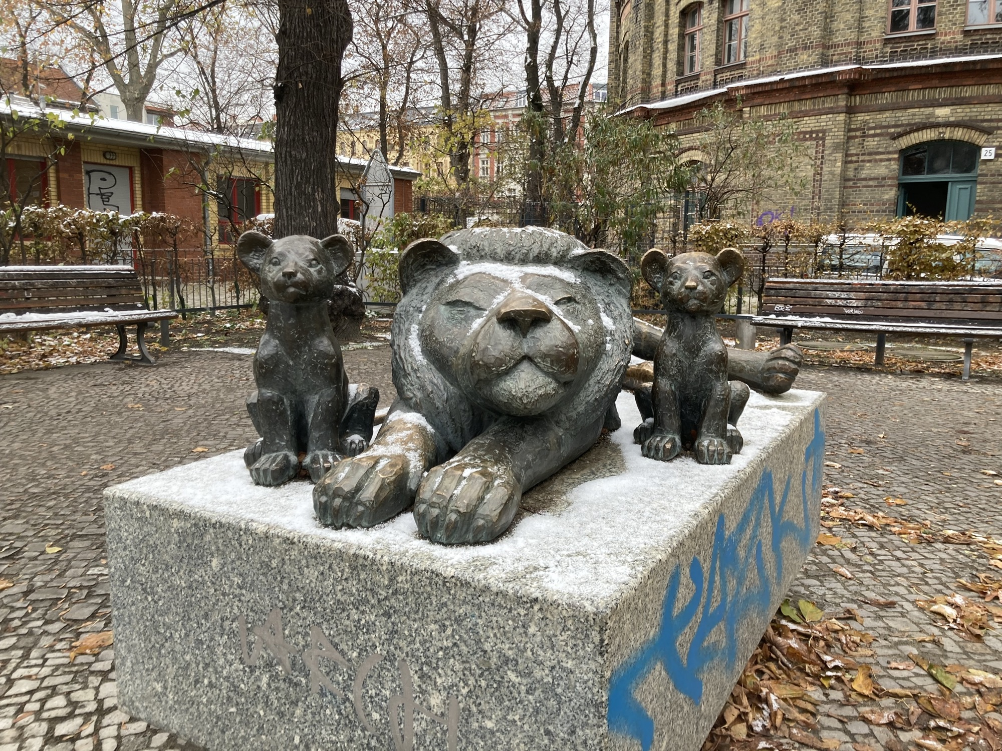 Metall-Skulptur eines auf der Seite liegenden, erwachsenen Löwen, flankiert von zwei sitzenden Löwenkindern, die Skulptur ist von einem dünnen Schneefilm bedeckt