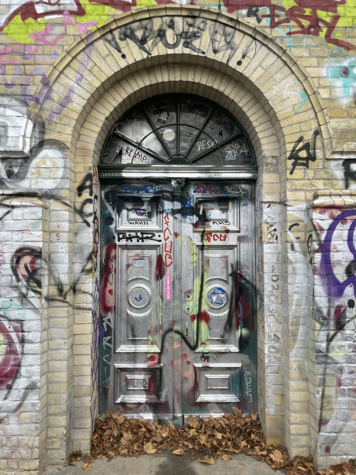 Türbereich des schmäleren Wasserturms auf dem Hügel, die Tür beinhaltet oben einen halbkreisförmigen Abschluss, der von einem Bogen aus Ziegeln eingefasst ist, sowohl Tür als auch die Wände drumherum sind mit verschiedenfarbigem Graffiti bedeckt, unten vor der Tür liegt eine Schicht Laub