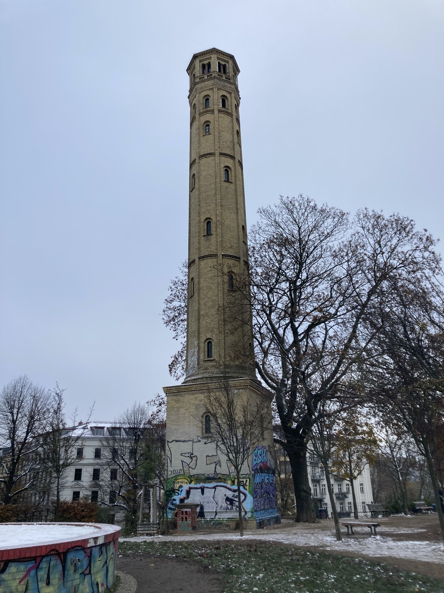 Gesamtansicht des schmäleren Wasserturms auf dem Hügel, eingefasst von blattlosen Bäumen, der untere Bereich des Turms ist mit Graffiti bedeckt