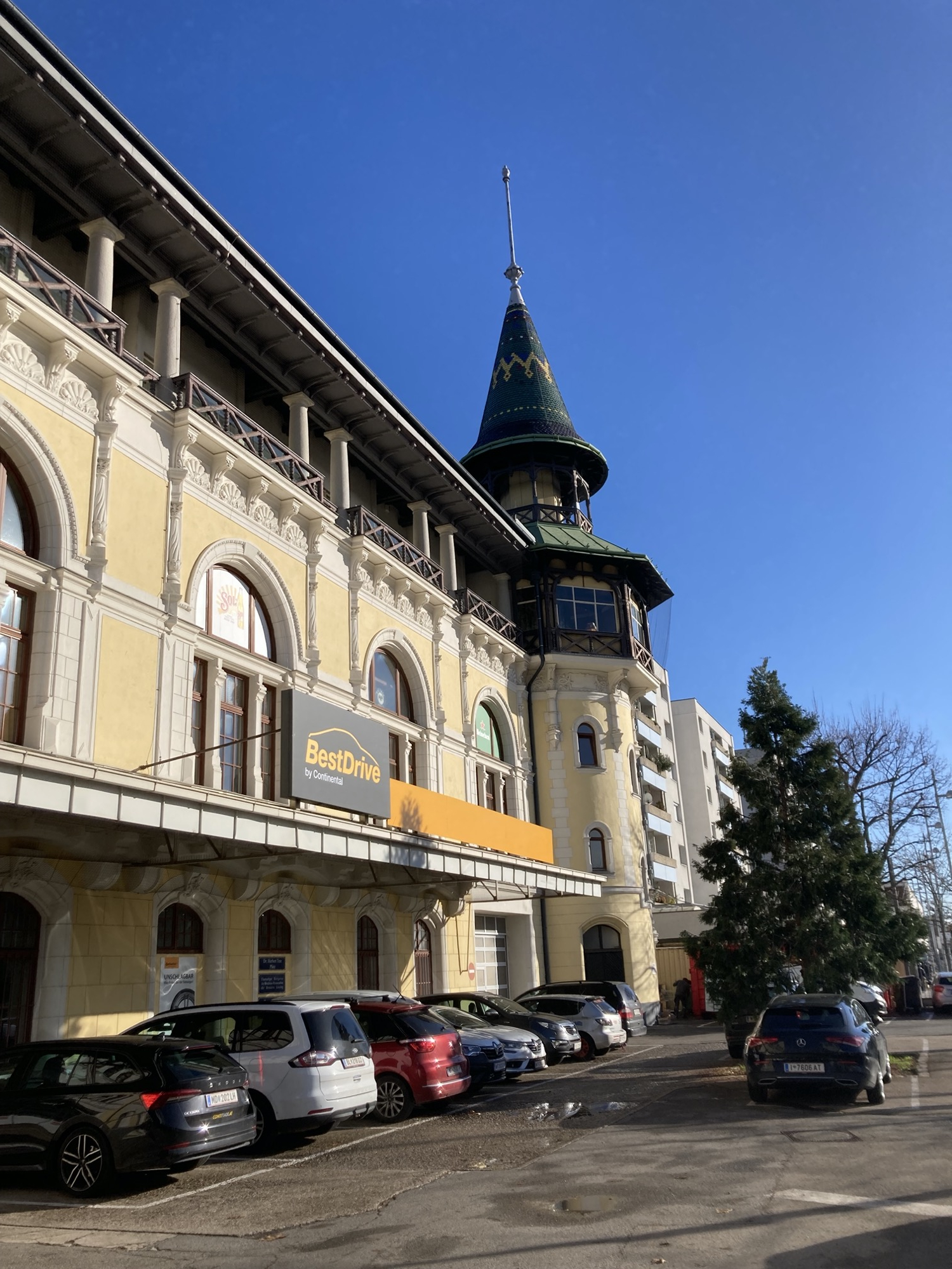 Vorderseite des ehemaligen Gebäudes der Brauerei Liesing, gelbe Fassade mit weißen Verzierungen und einem Eckturm mit Erker, Balkon und dekoriertem Dach