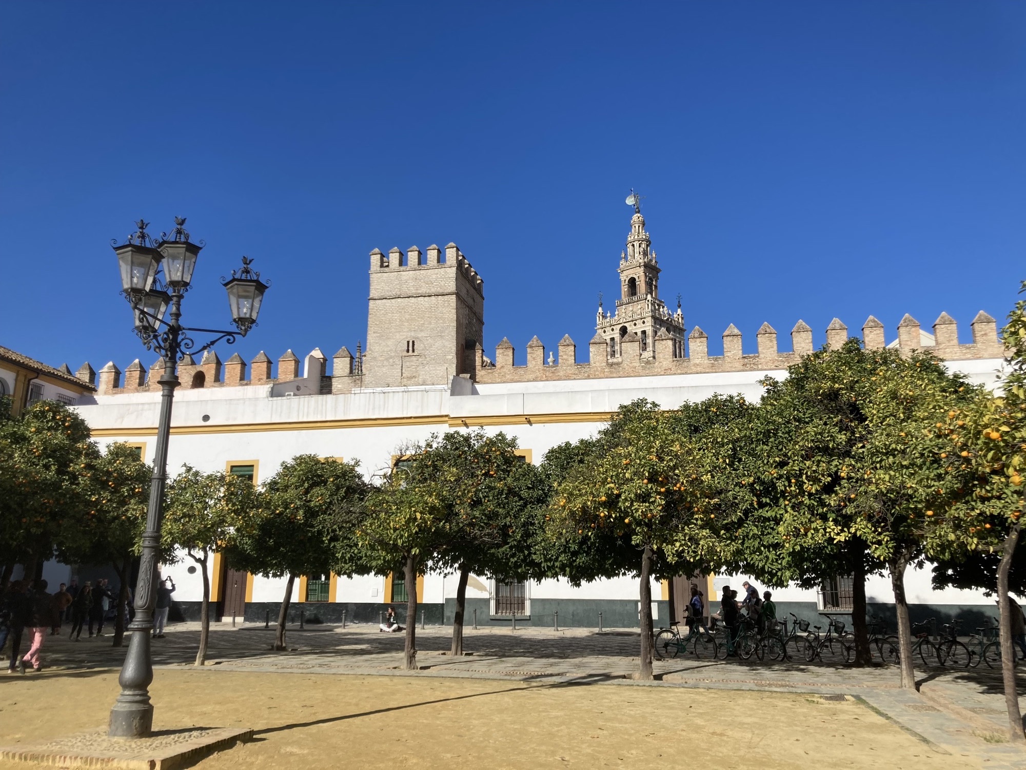 Orangenhof des Alcázar von Sevilla, im Vordergrund eine vierköpfige Lampe und mehrere Orangenbäume, dahinter die Mauern des Alcázars, geschmückt mit Rauchfang-ähnlichen Spitzen, dahinter ragt der Glockenturm Giralda über die Mauer hinaus