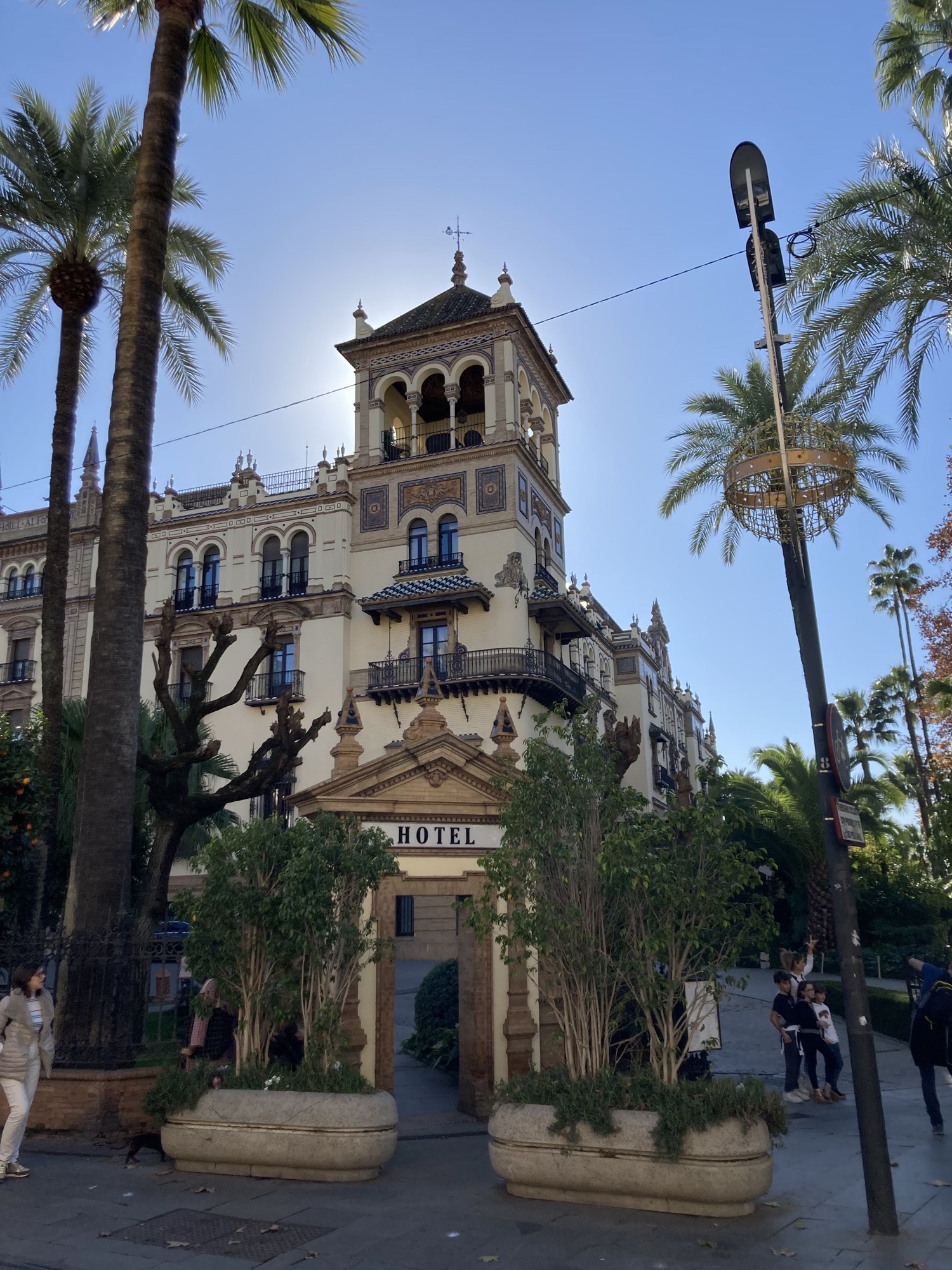 Hotel Alfonso XIII, Eckturm des Hauses mit unzähligen Verzierungen aus Keramikmosaiken, schmiedeeisernen Gittern, Rundbögen und Ziergiebeln, direkt hinter dem Turm ist die Sonne verdeckt und verleiht dem Turm somit ein Glühen, links und rechts Palmen, sowie eine Straßenlaterne mit Weihnachtsdekoration