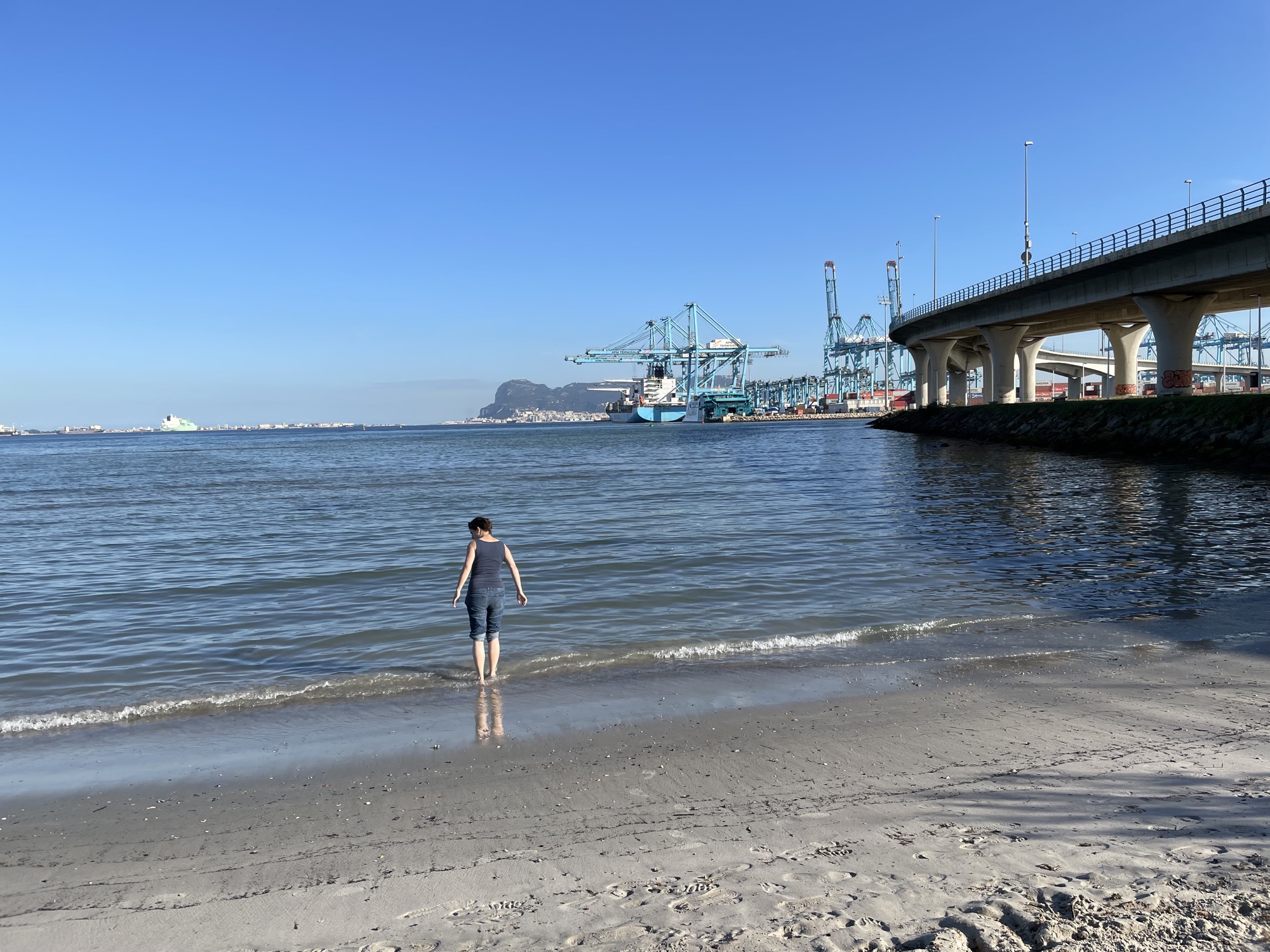 am Strand von Algeciras, eine Person steht mit bis zu den Knien aufgekrempelten Hosen im seichten Wasser, rechts führt eine Brücke zum Containerhafen, ganz hinten mittig im Bild ist der Felsen von Gibraltar zu sehen