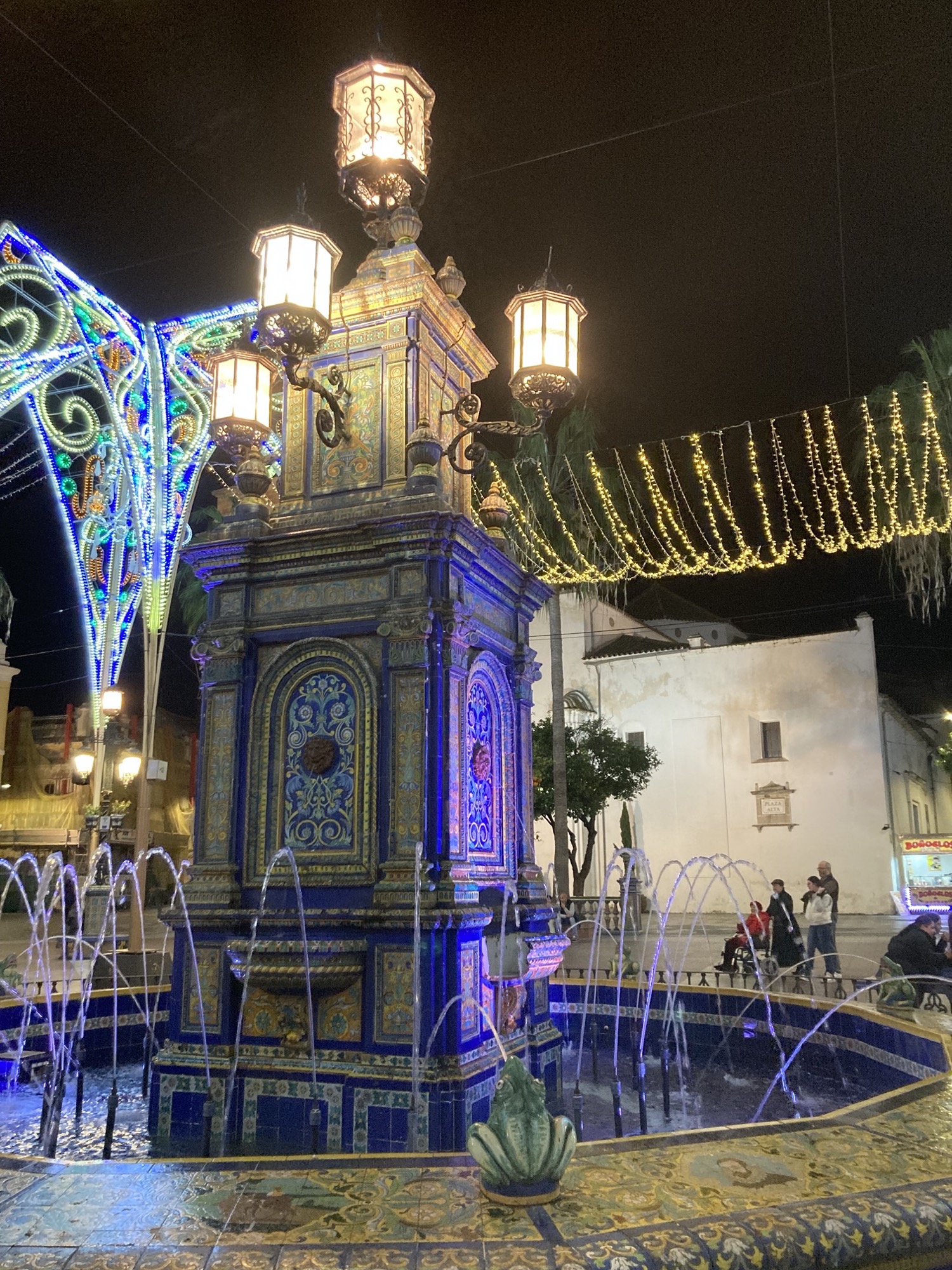 der Brunnen auf dem Hauptplatz von Algeciras ist mit einem detaillierten Mosaik geschmückt, die Säule in der Mitte ist mit vier Lampen gekrönt, rundherum spucken Froschskulpturen Wasserfontänen in den Brunnen, hinter dem Brunnen ist Weihnachtsbeleuchtung zu sehen