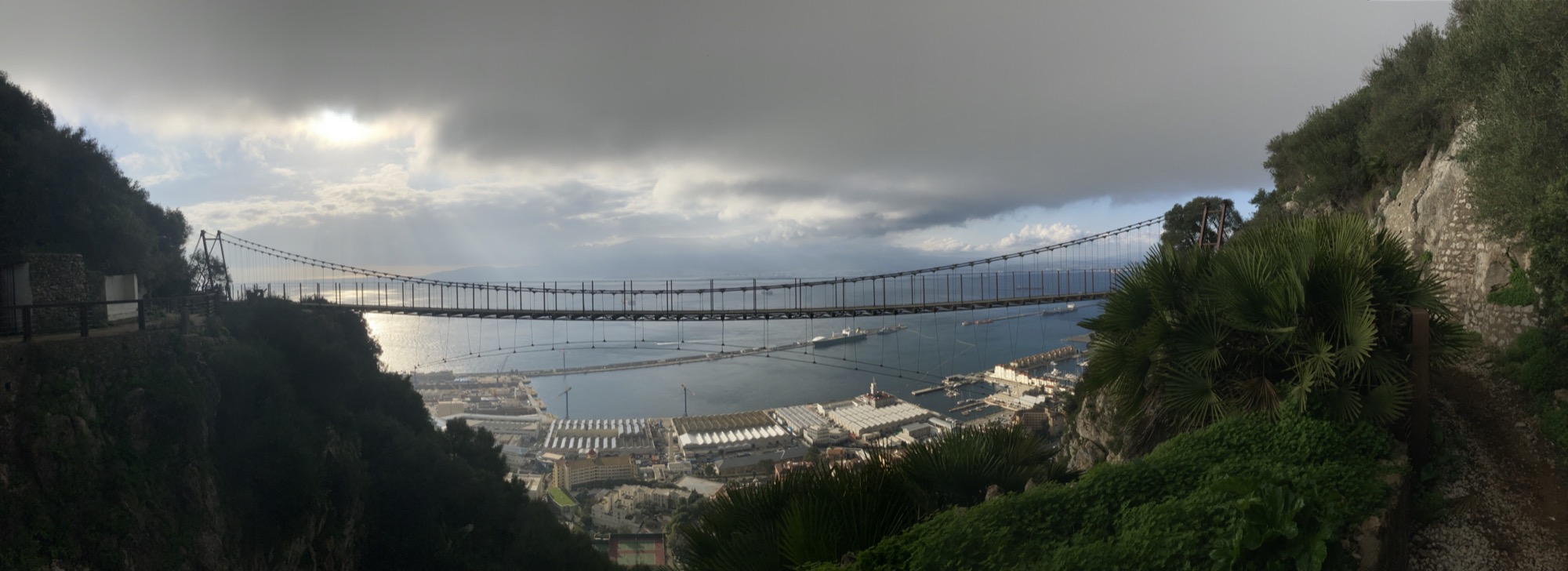 Panoramaaufnahme der Hängebrücke in voller Länge, dahinter bzw. darunter sind Industriegebäude sowie ankernde Frachtschiffe im Hafen von Gibraltar zu sehen