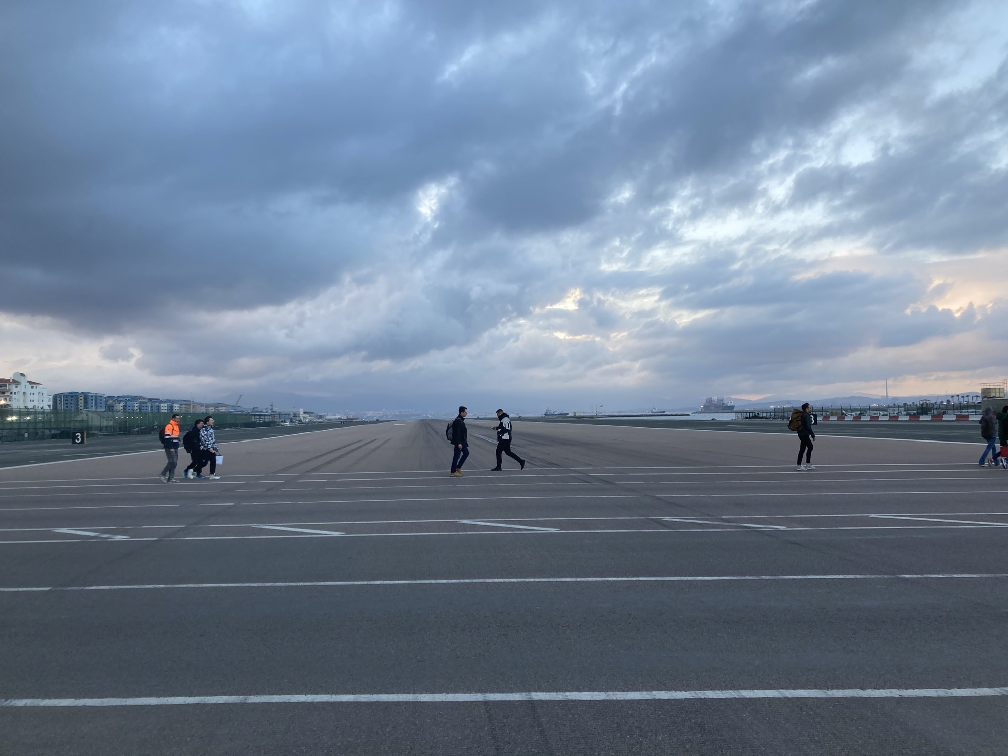 Blick vom Gehweg auf das Flugfeld des Flughafens Gibraltar, auf dem Fußweg auf der anderen Seite der Straße sind mehrere Personen zu sehen, die in unterschiedliche Richtungen das Flugfeld überqueren. Die Dämmerung hat bereits eingesetzt, die Wolkendecke hat eine deutlich bläuliche Färbung angenommen