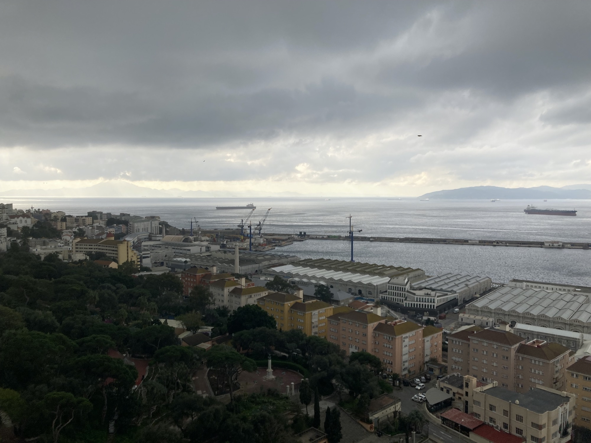 Ausblick aus der Kabine der Seilbahn, im Vordergrund sind ein Park, Wohnhäuser und Industriegebäude am Hafen von Gibraltar zu sehen, zwei Frachtschiffe kreuzen die Bucht, am Horizont ist rechts die südlichste Spitze von Spanien südlich von Algeciras zu sehen, weiter links im Hintergrund weiter entfernte Berge, dies ist bereits Afrika, über all dem schwebt ein düsterer grauer Wolkenhimmel, der nur am Horizont etwas aufreißt