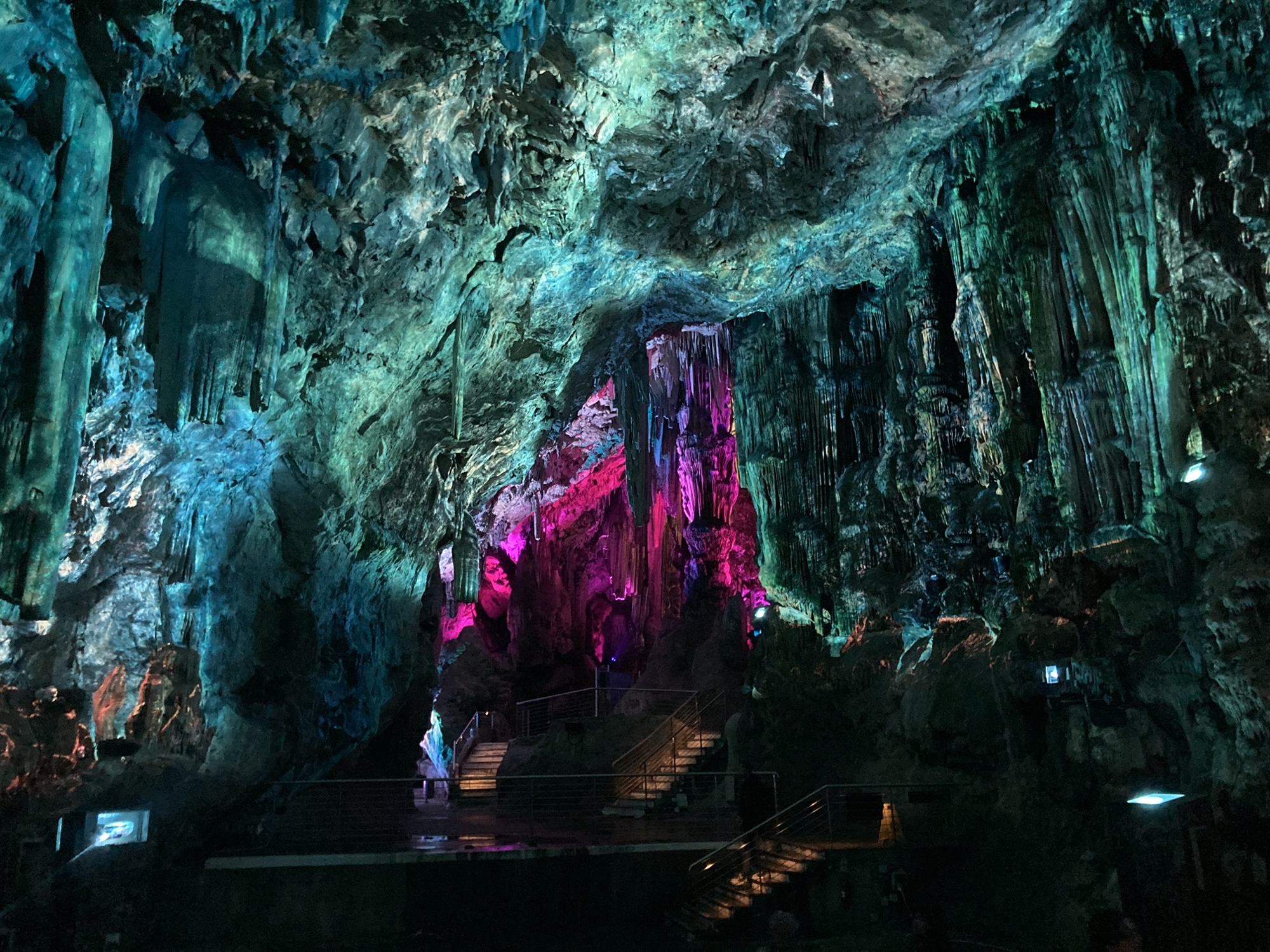 Auditorium in der großen Halle der Höhle, die Stalagtiten an der Decke sind hell in blau-grün beleuchtet, sodass sie wie fließendes Wasser wirken, mittig im Hintergrund leuchtet der Ausgang aus dem Auditorium in pink-violett