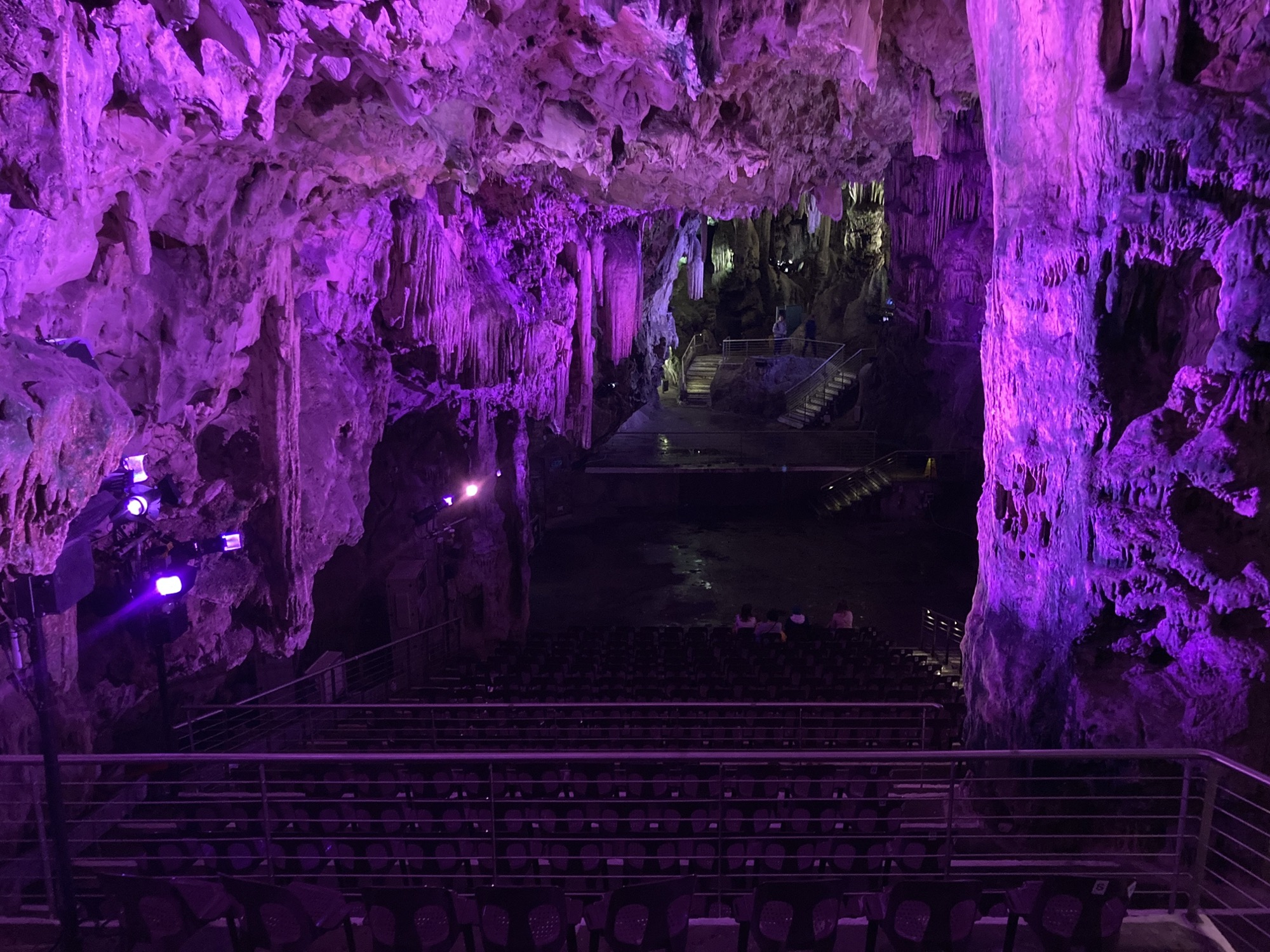Auditorium in der großen Halle der Höhle, Blick aus dem Zuschauerraum Richtung Bühne, das gesamte Bild ist in violettes Licht getaucht, in der ersten Reihe sitzen vier Personen mit dem Rücken zur Kamera, auf dem Bühneboden spiegelt sich das bunte Licht in Wasser, das von der Decke getropft ist