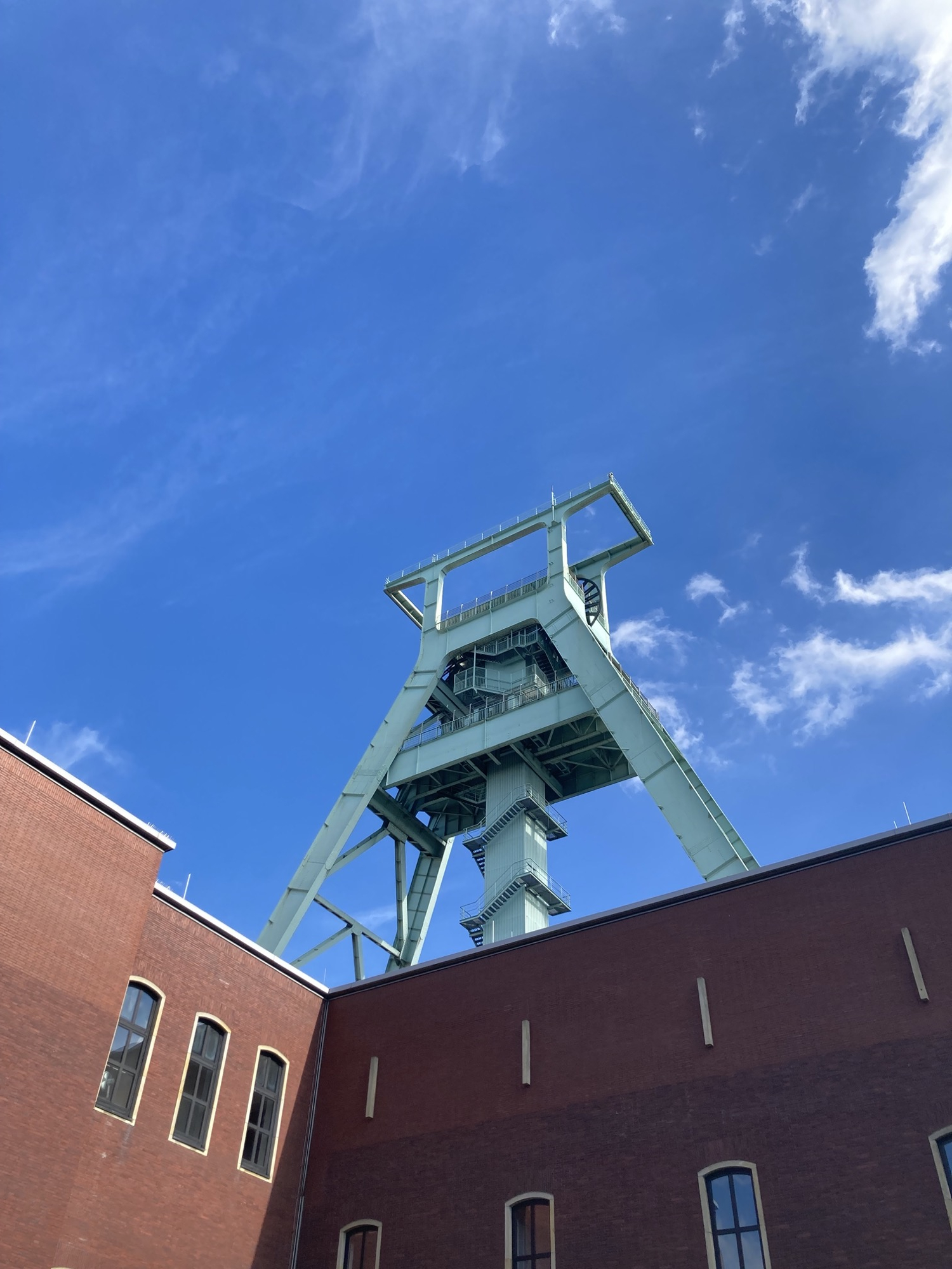 Deutsches Bergbau Museum, der Förderturm aus helltürkisem Stahl überragt das rote Ziegelgebäude vor blauem Himmel, in der Mitte des Förderturms windet sich eine Stiege den Mittelpfosten hinauf