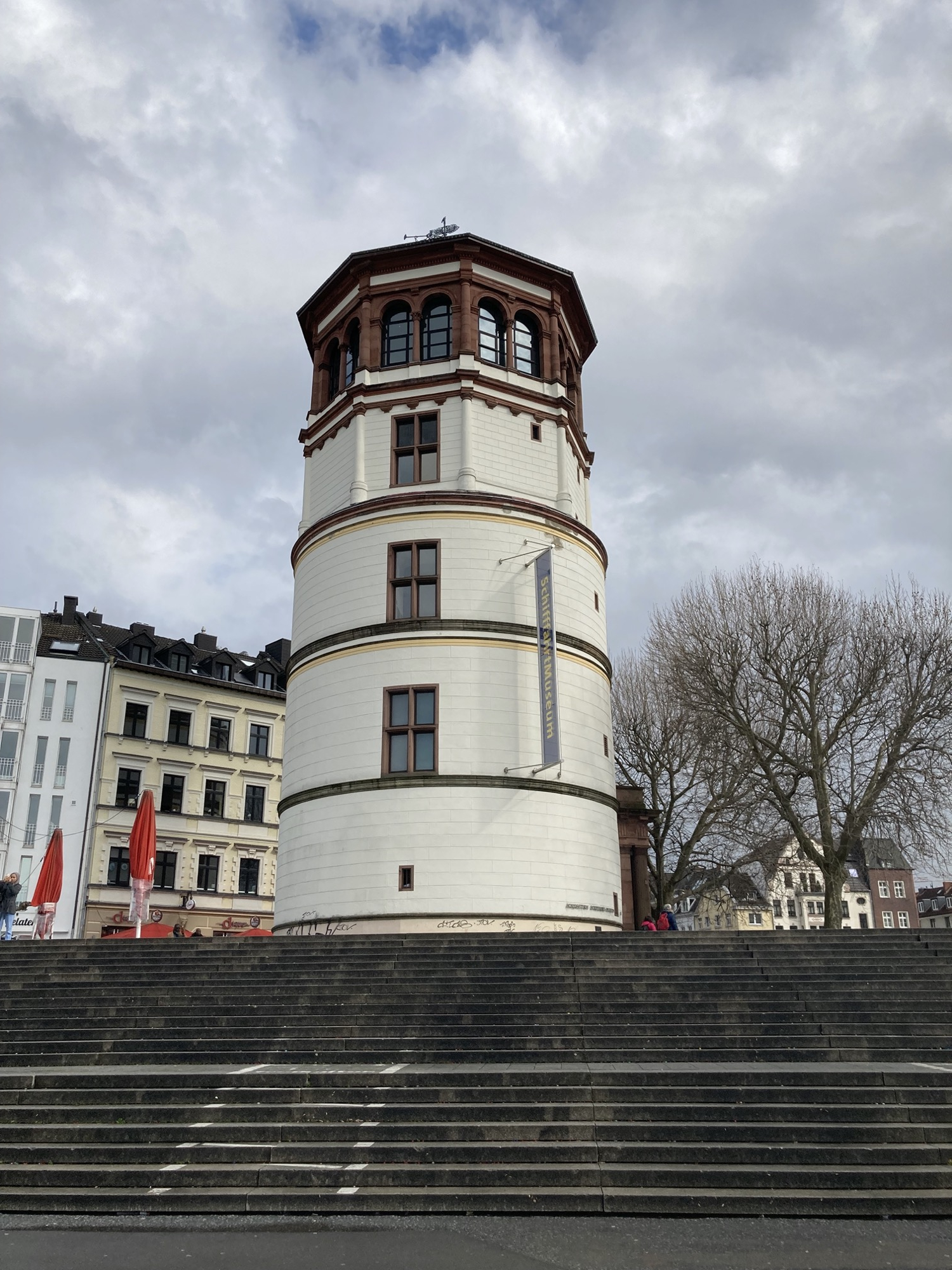 Schloss-Turm am Burgplatz von der Rhein-Promenade aus gesehen, der Turm hat fünf Stockwerke, die unteren sind hell bemalt, ein Transparent weist auf das Schifffahrtsmuseum hin, das oberste Stockwerk ist aus braunen Ziegeln, hat rundherum Fenster und lässt den Turm an einen Leuchtturm erinnern