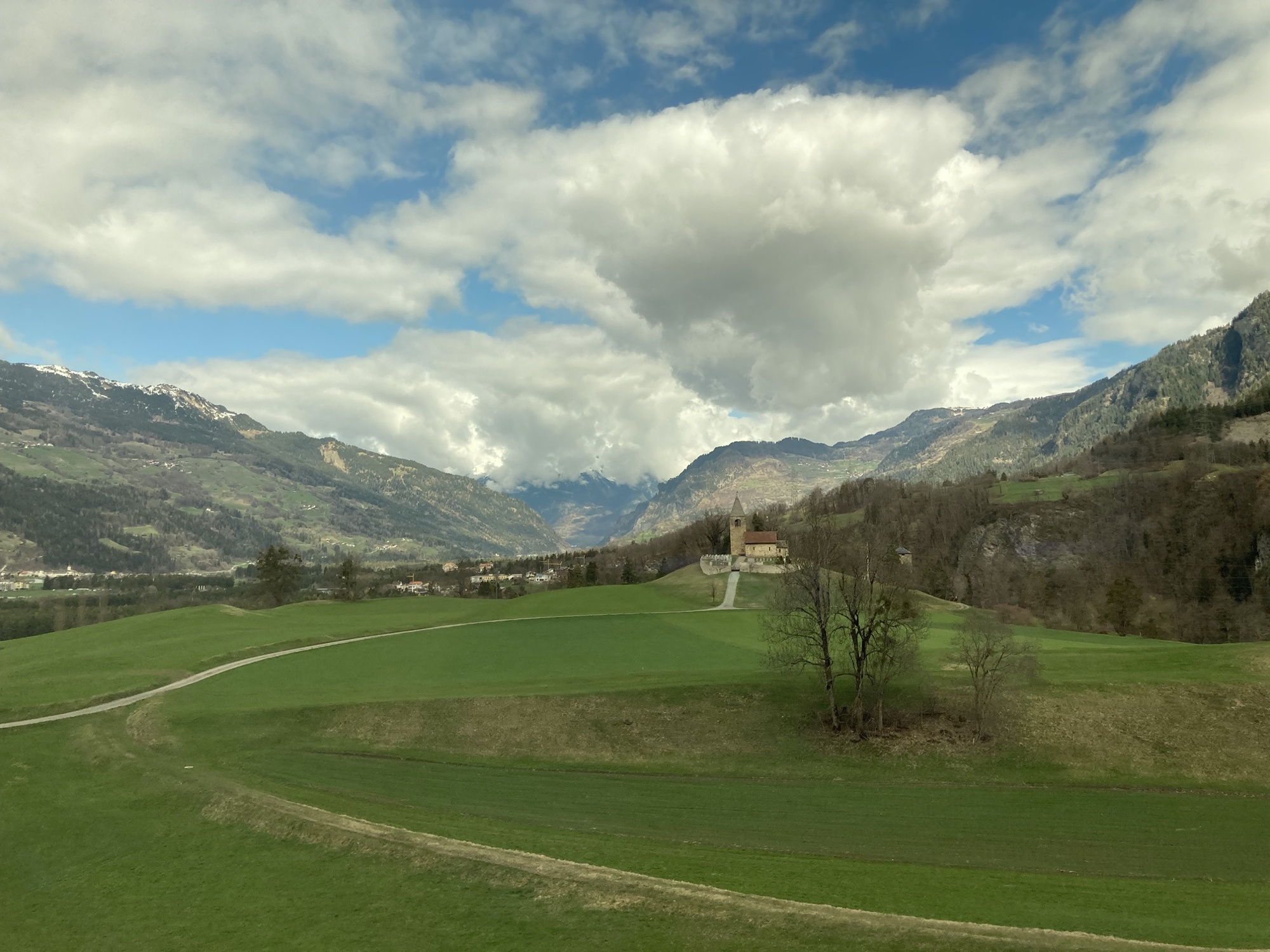 Schweizer Landschaft, Feldweg, der zu einer Kirche auf einem Hügel führt, Berge mit schneebedeckten Spitzen, Wolken vor blauem Himmel