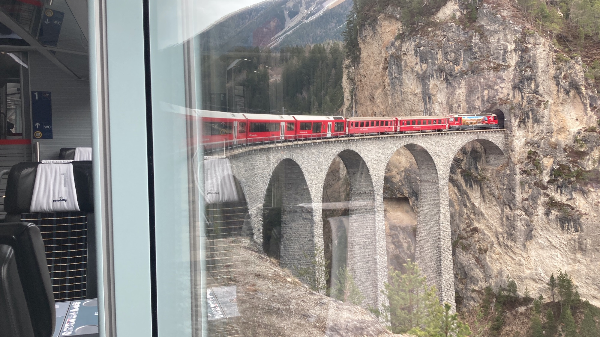 Blick aus dem Zugfenster des letzten Waggons, der Zug fährt gerade in eine Kurve, die Lok und die vorderen Waggons fahren auf einem sehr hohen Viadukt über eine Schlucht, die Lok fährt im nächsten Moment in einen Tunnel ein, der an das Viadukt anschließt