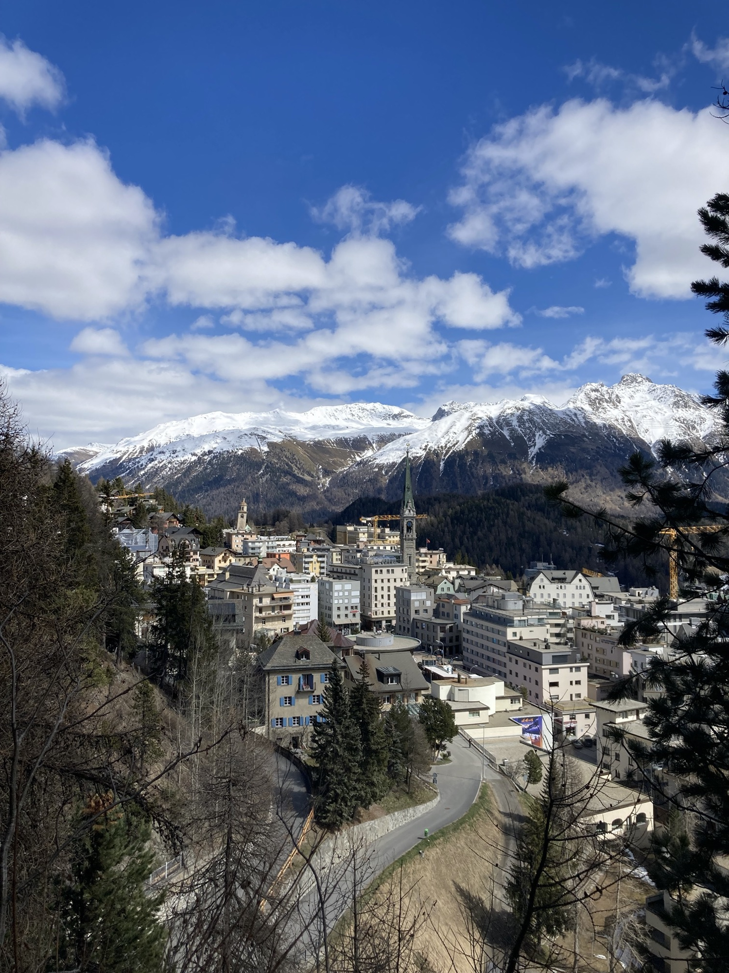 Überblick über die Stadt St. Moritz, der Kirchturm, der schiefe Turm und ein Baukran überragen die vielen Hotelgebäude, im Hintergrund eine Bergkette mit schneebedeckten Gipfeln und leicht bewölkter Himmel