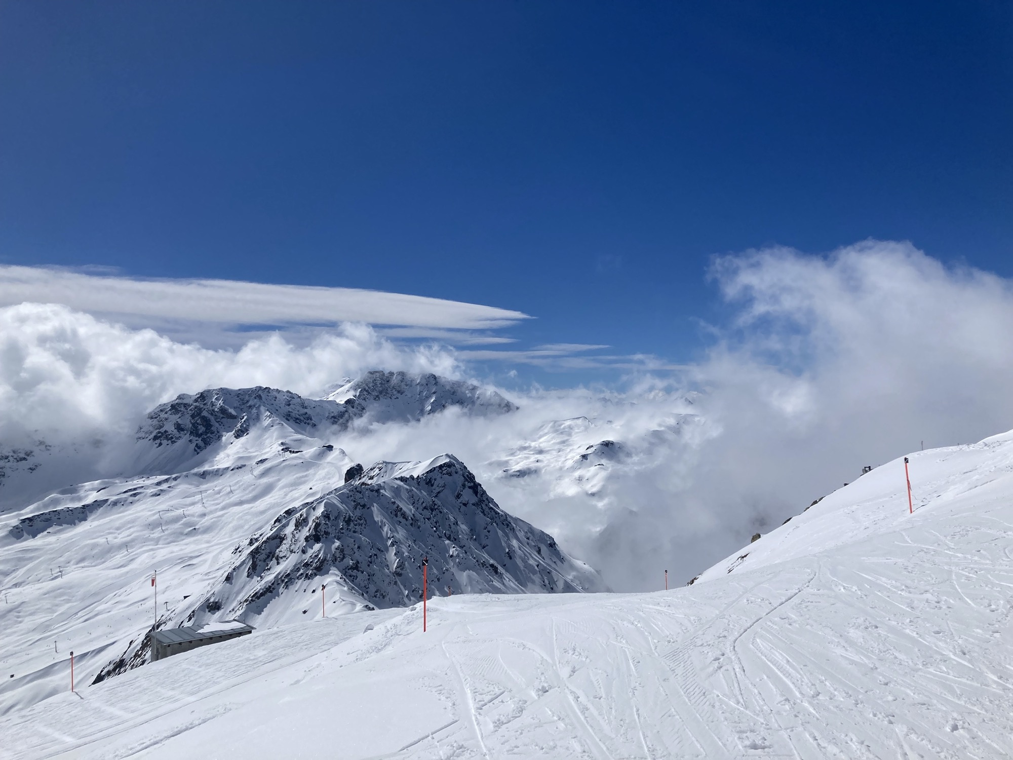 Postkarten-taugliches Bergpanorama, von Wolken umschmeichelte Berggipfel ragen zwischen Skipisten in den tiefblauen Himmel