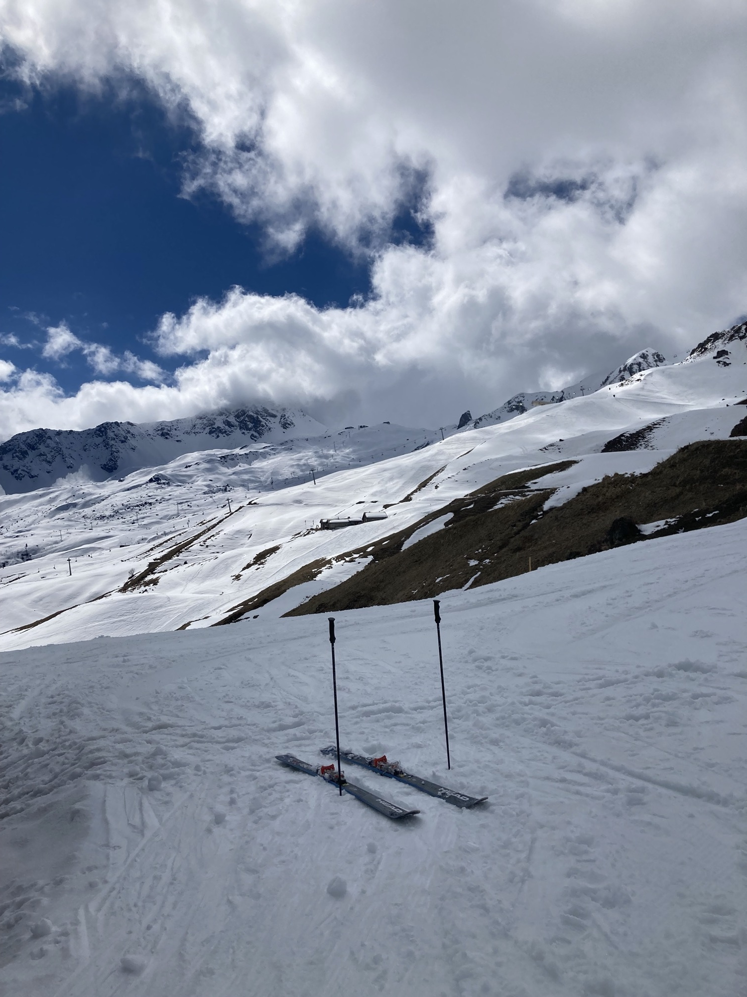 Skigebiet-Idylle, im Vordergrund ein geparktes Paar Ski mit daneben in den Schnee gesteckten Skistöcken, im Hintergrund sonnenbeschiene Skipisten unter blauem Himmel
