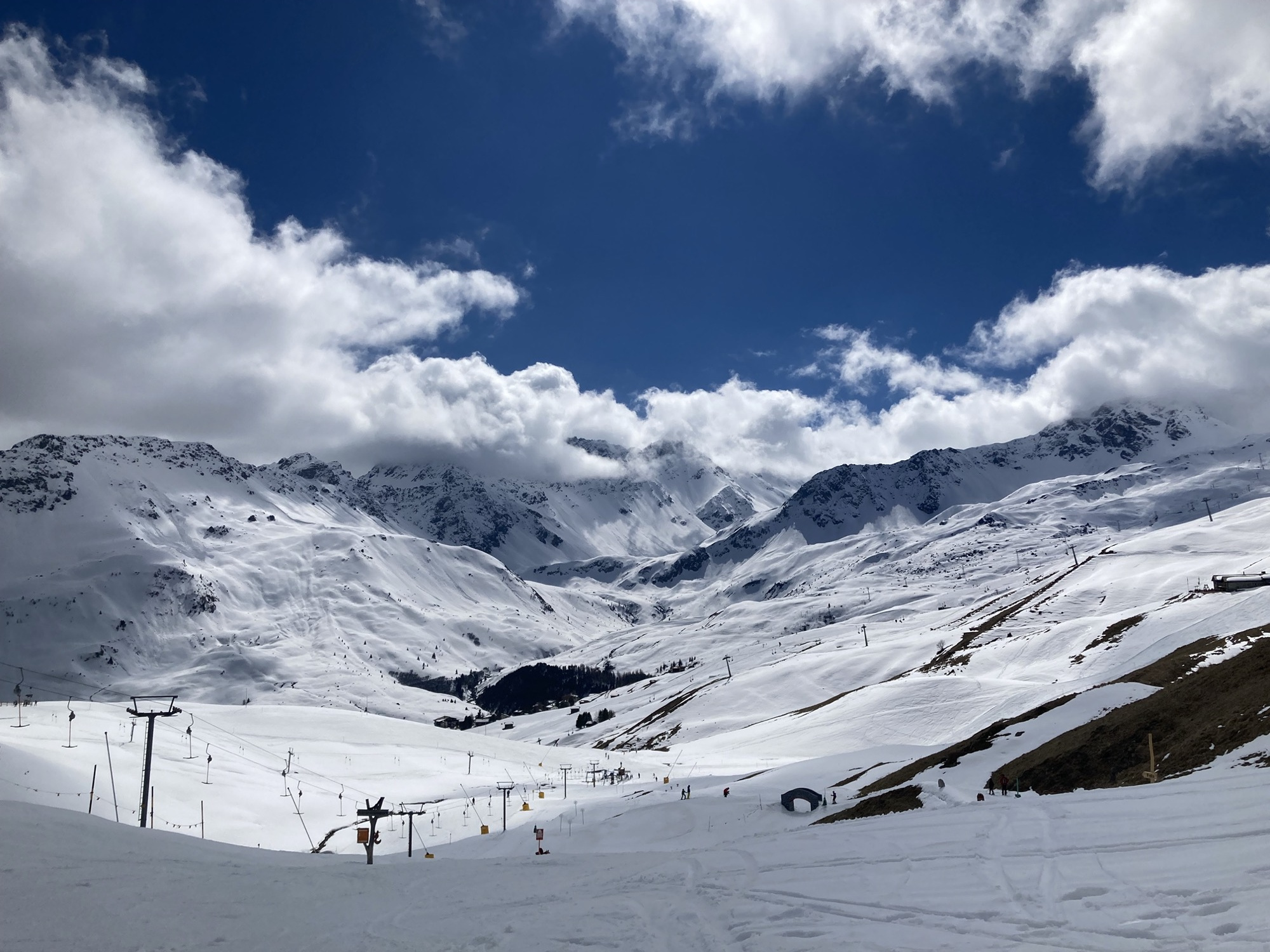 Skigebiet-Idylle, Blick in ein Tal mit mehreren Liftanlagen zwischen Skipisten, im Hintergrund wolkenverhangene Berggipfel vor strahlend blauem Himmel