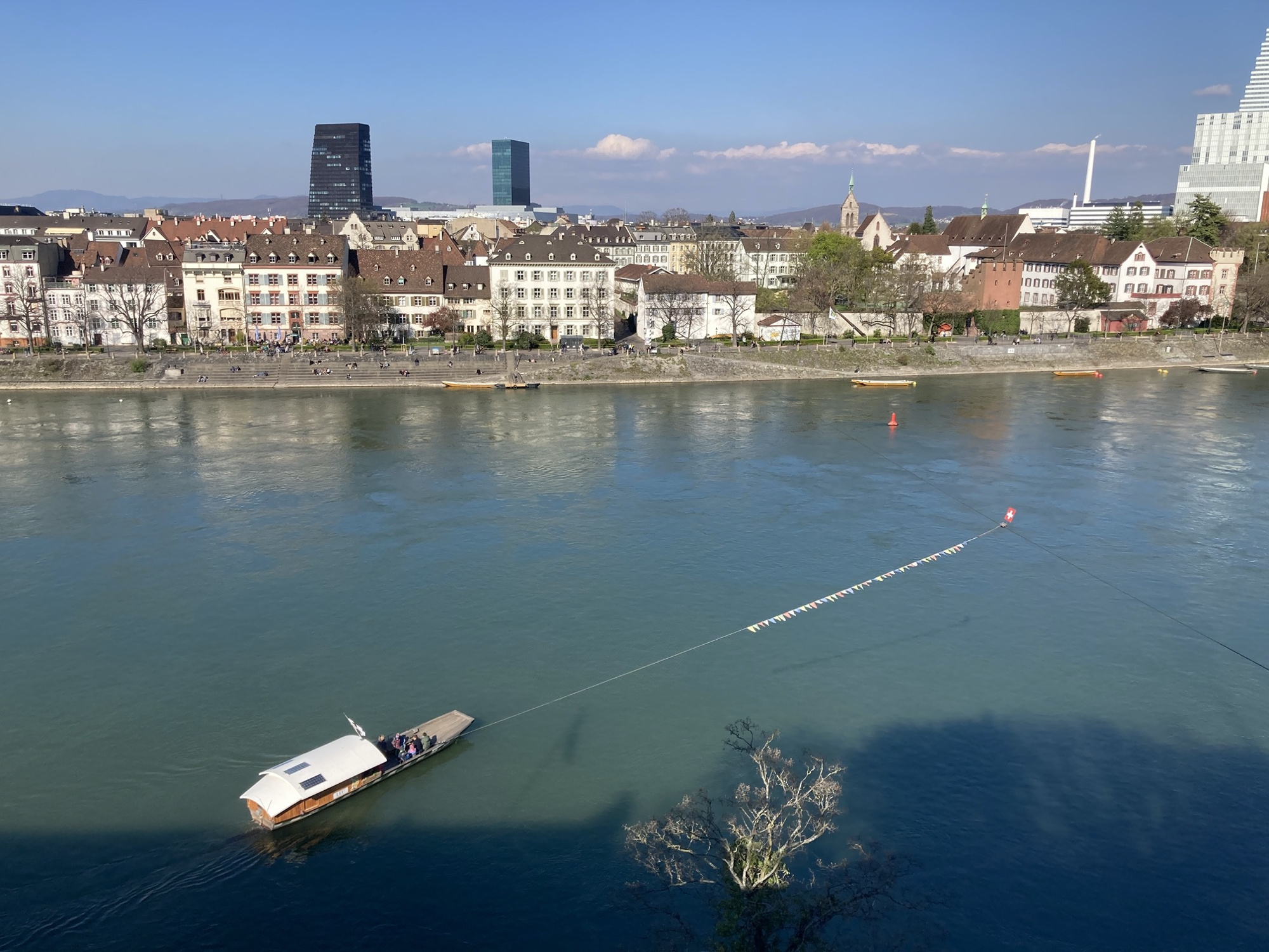 Fähre, die an einem Seil über den Rhein gezogen wird, im Hintergrund der Stadtteil Leu mit Uferpromenade, alten Häusern und zwei vereinzelten Wolkenkratzern