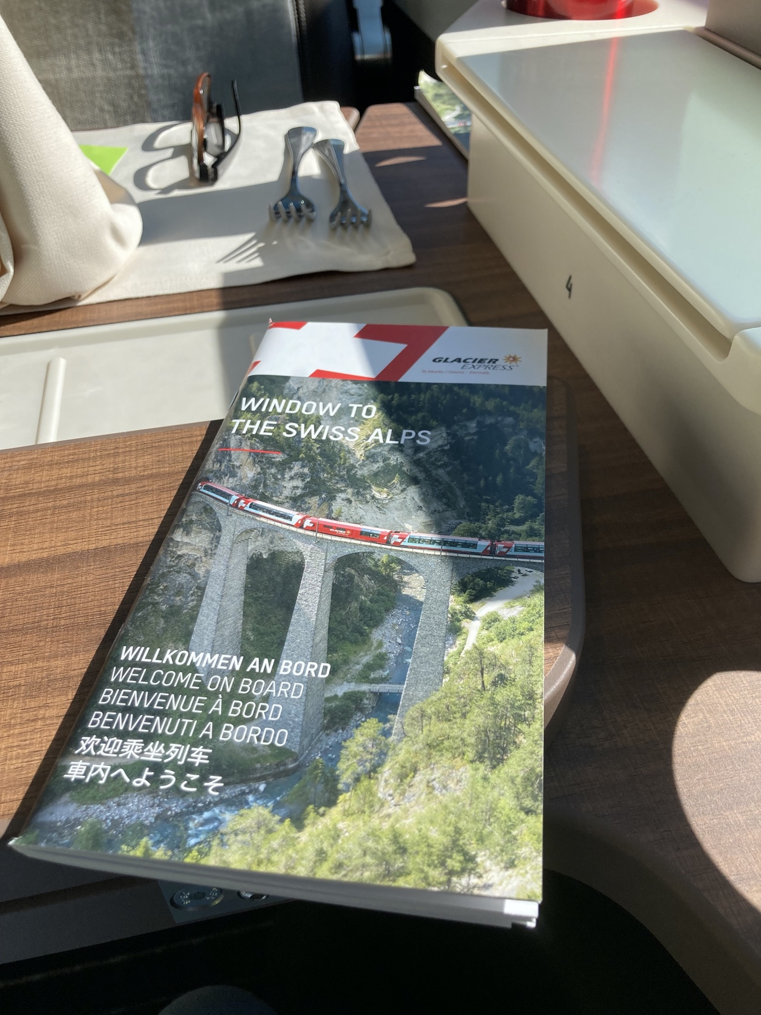 Broschüre zum Glacier Express mit dem Text „Window to the Swiss Alps“ und „Willkommen an Bord“ in 6 Sprachen, ein Foto eines Zuges auf einem hohen Viadukt, die Broschüre liegt auf einem Tisch im Waggon, dahinter Serviette, zwei Gabeln und eine Brille