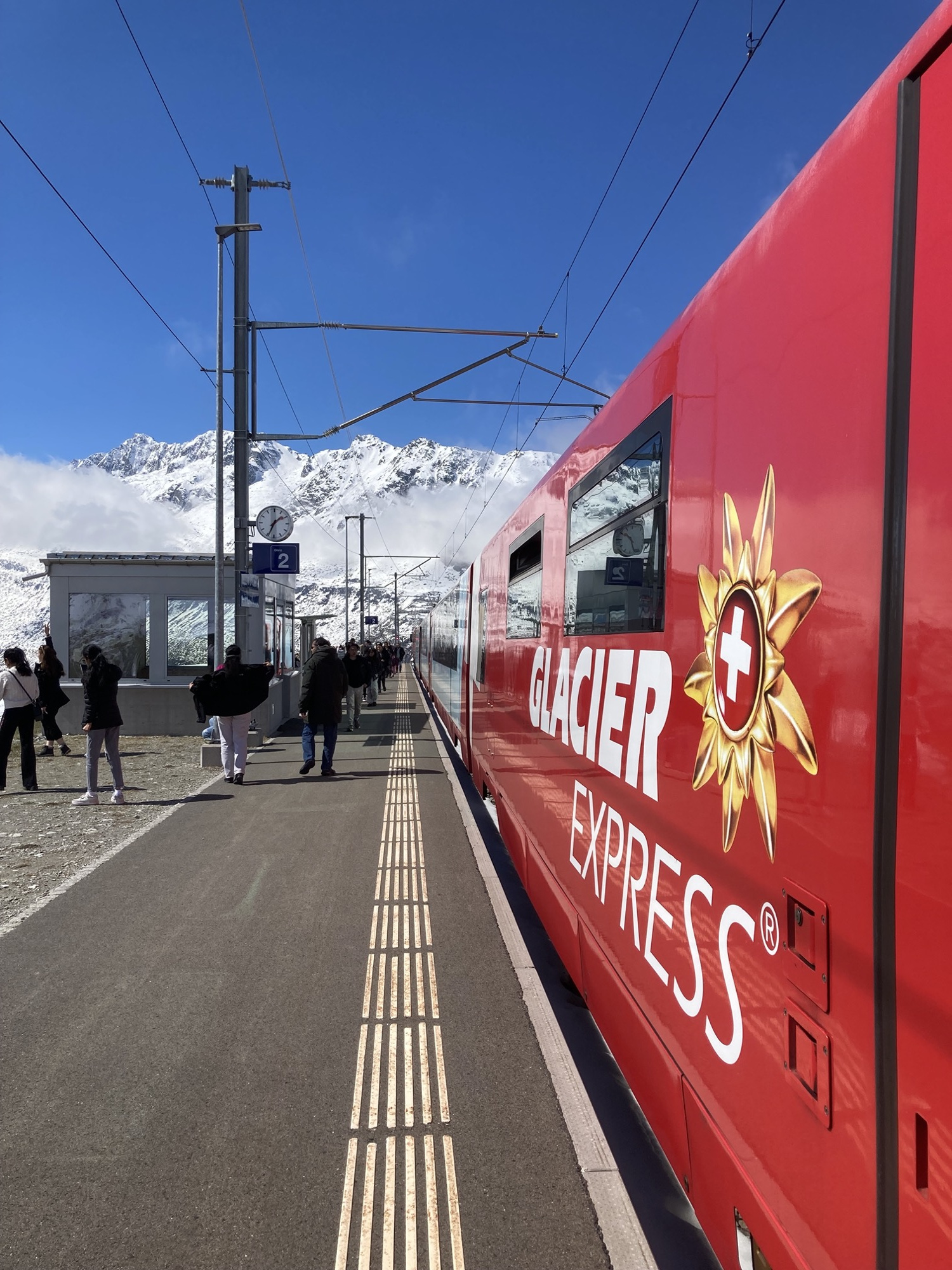 Bahnsteigblick, rechts der rote Waggon mit der Aufschrift „Glacier Express“, im Hintergrund ein Bergpanorama, auf dem Bahnsteig einige Menschen, die von der Kamera wegschauen auf die Berge rundherum