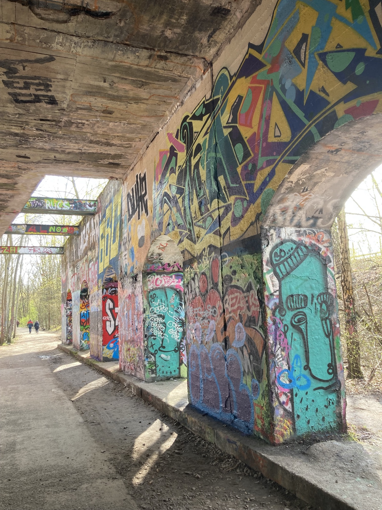 Weg unter einer Eisenbahnbrücke, die Pfeiler der Brücke sind mit Graffiti bemalt, zwischen den Pfeilern der Brücke scheint Sonnenlicht auf den Weg