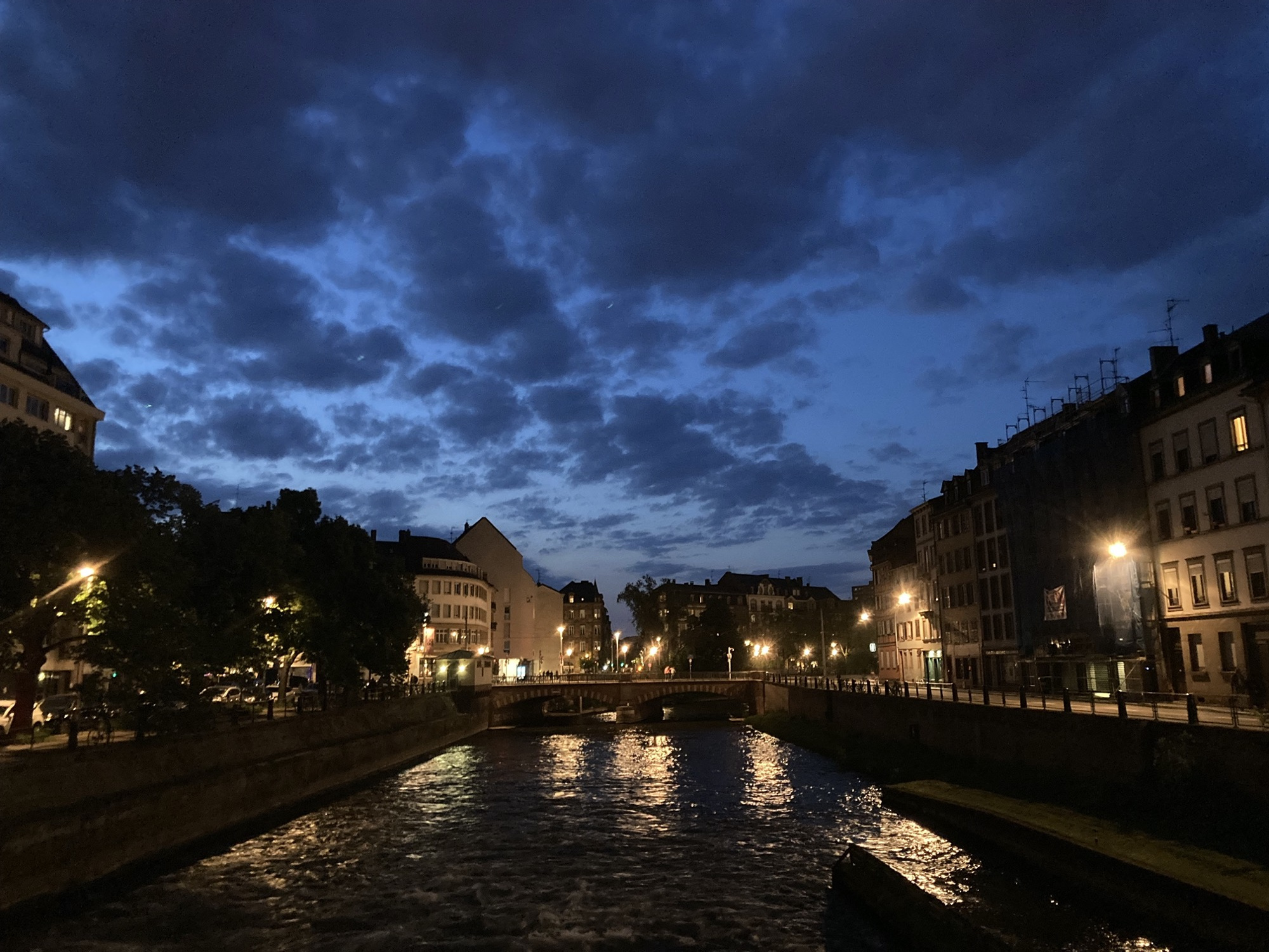 Brücke über einen Fluss, im Fluss spiegeln sich die Lichter der umliegenden Gebäude, mittig im Hintergrund ist der gerade noch dunkelblaue Himmel leicht bewölkt