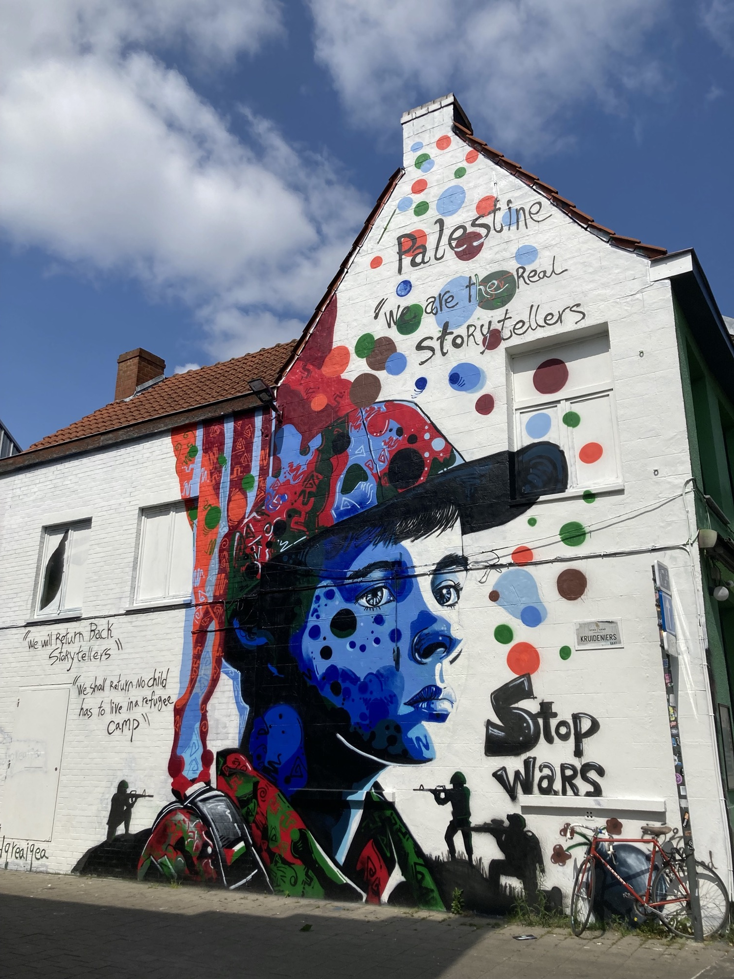 Graffiti an der weißen Wand eines Hauses, ein Porträt einer Person mit Schirmkappe, die von kleinen Soldaten mit Waffen bedroht wird, Text drumherum: „Palestine“, „we are the real storytellers“, „stop wars“
