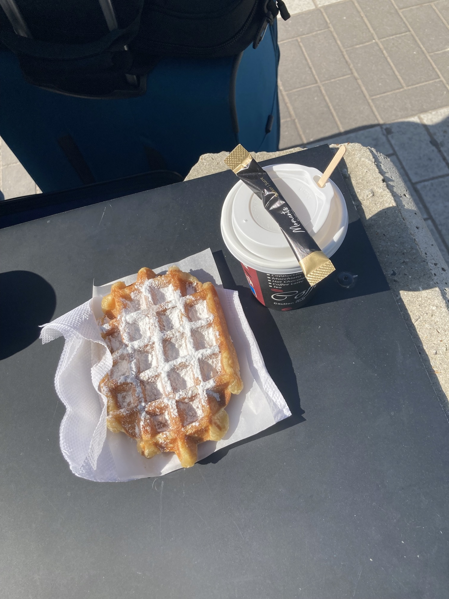 Belgische Waffel mit Zucker, daneben ein Kaffee im Take-away-Becher mit Holzstäbchen und Zucker
