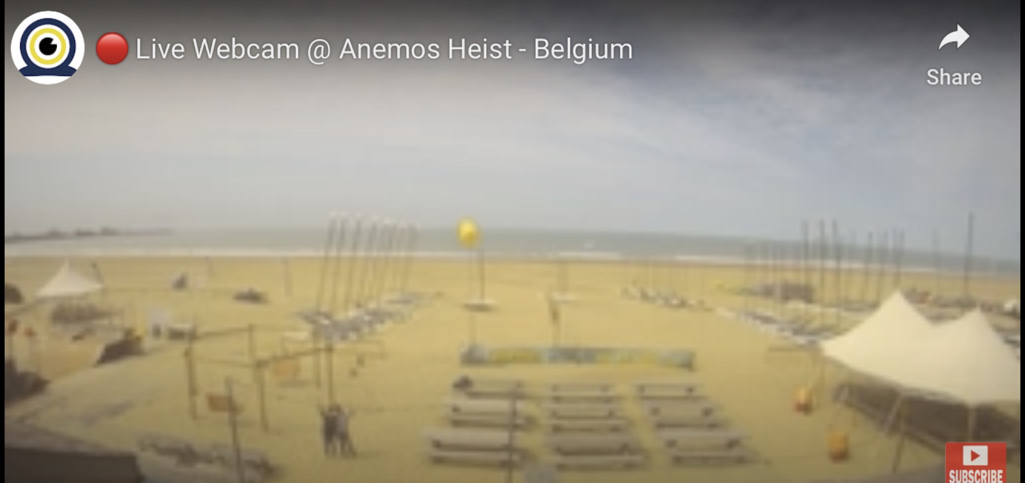 Live Webcam @ Anemos Heist Belgium, verschwommener Screenshot eines menschenleeren Strandes mit Zelten, Booten und Bänken, in der linken Hälfte des Bildes zwei Personen, die nebeneinander stehen und ihre Arme hochhalten