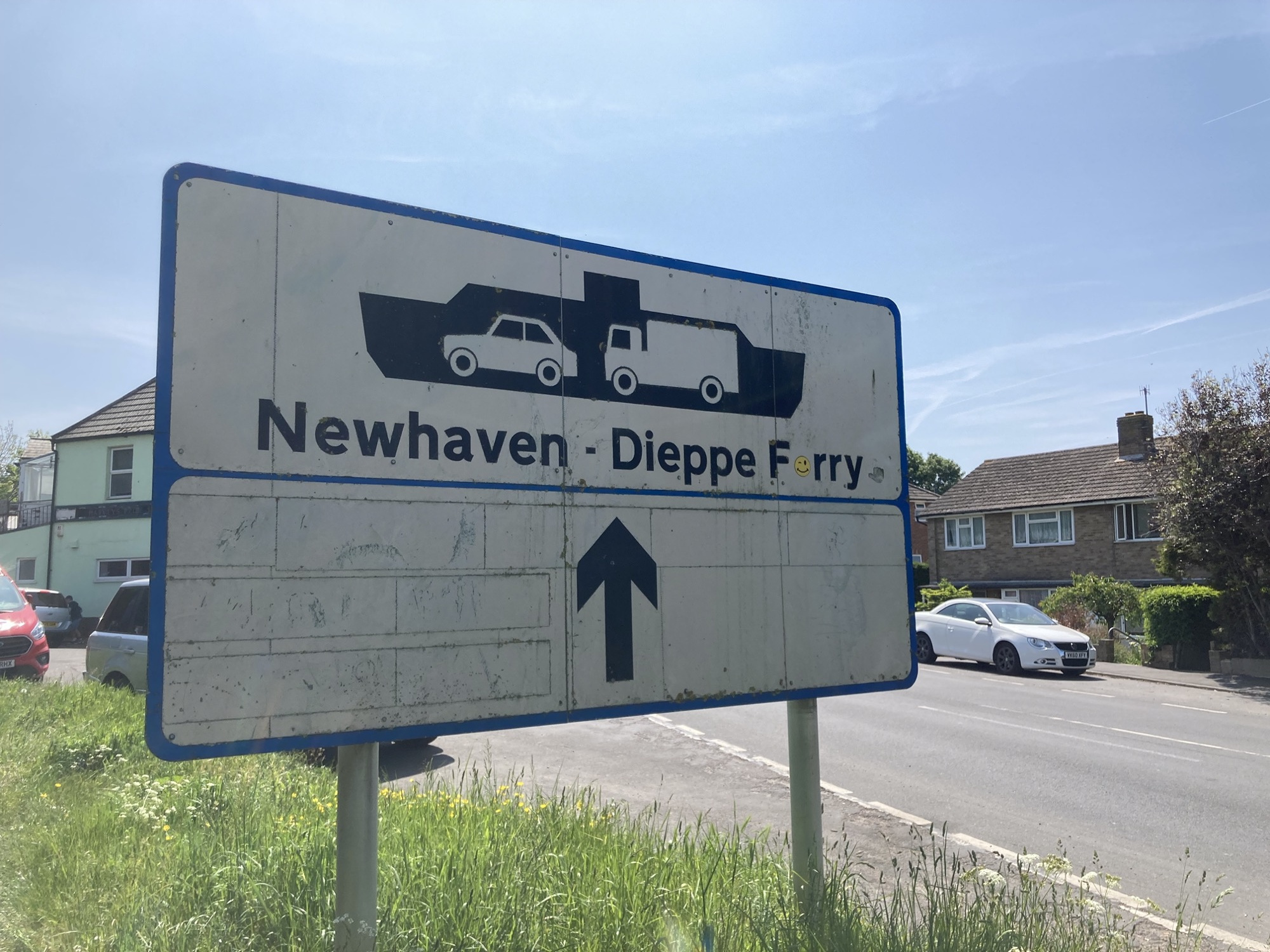 Straßenschild, das die Richtung zur Fähre „Newhaven – Dieppe“ anzeigt, über dem Text ist ein Piktogramm eines Fährschiffes mit einem PKW und einem LKW im Schiffsbauch