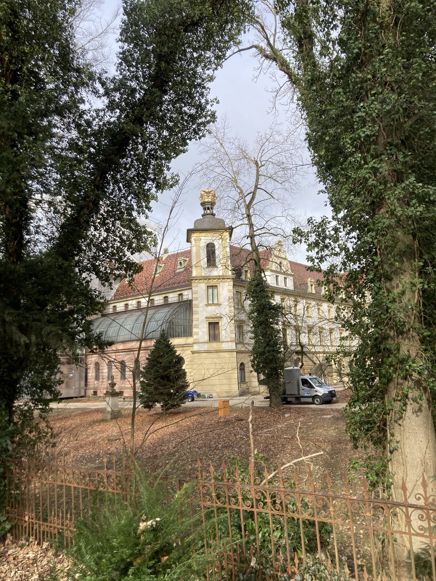 Schloss St. Emmeram Thurn und Taxis, im Vordergrund ein Zaun und zwei Bäume an den Seiten des Bilds, sie umrahmen einen Eckturm eines imposanten Gebäudes mit einem halbrunden Glasdach links und einem rot geziegelten Dach, der Turm ist an der Spitze von einer goldenen Krone geschmückt