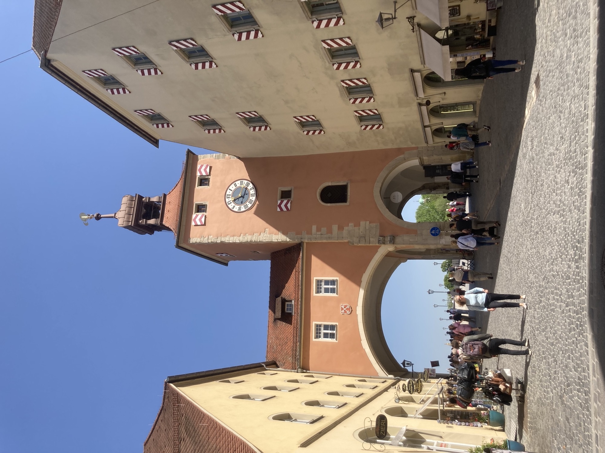 Brückturm in der Altstadt von Regensburg, links und rechts zwei Gebäude, dazwischen überspannen zwei Torbögen den Abstand, rechts ein Turm mit einer Uuhr daran, die Fenster haben rot-weiß-gestreifte Fensterläden