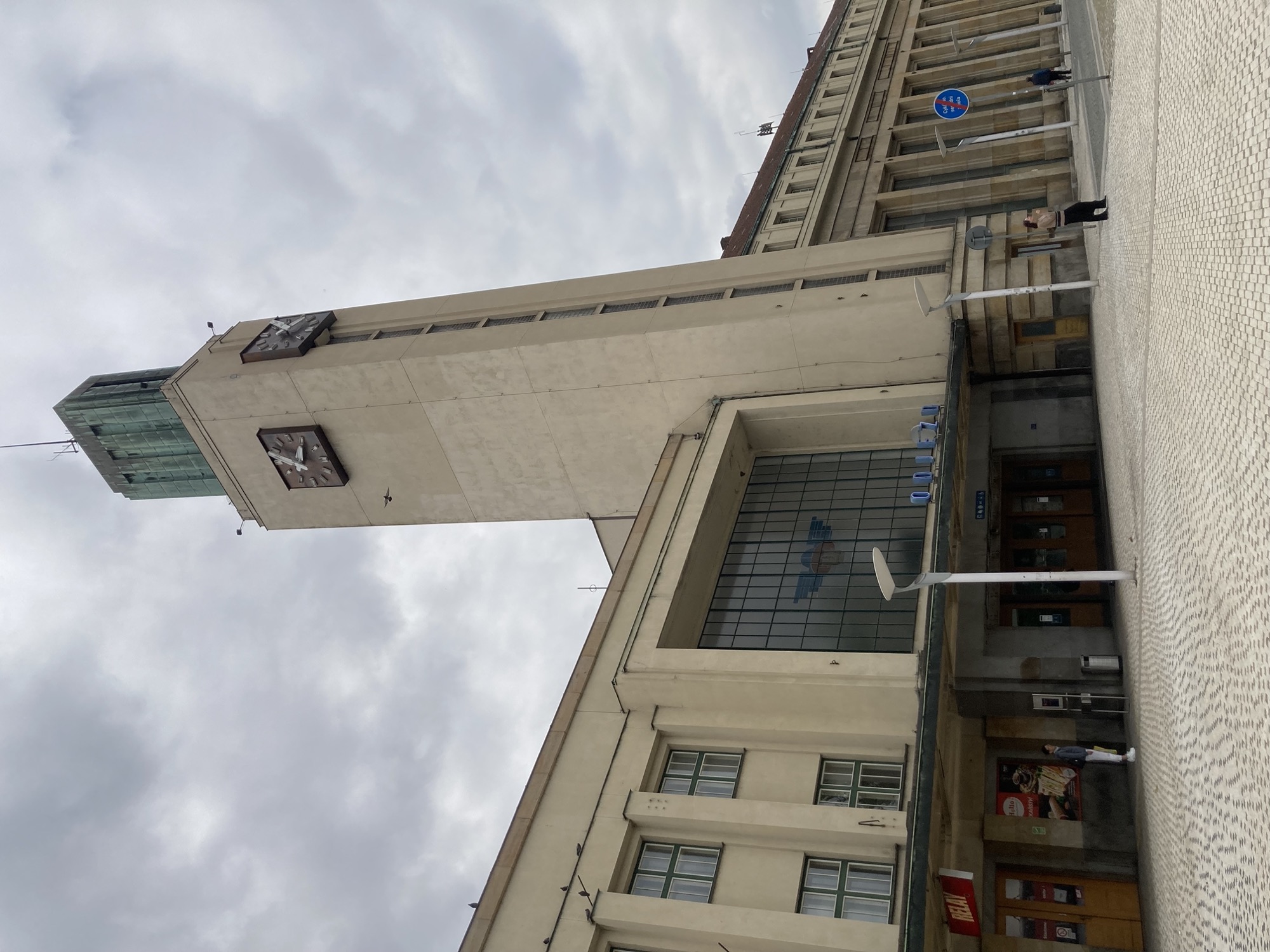 Bahnhofsgebäude, alles ist eckig, über dem Haupteingang ein großes Fenster mit einem Symbol, das an einen Vogel erinnert, daneben überragt ein rechteckiger Turm das Gebäude, oben ist an zwei Seiten eine Uhr zu sehen