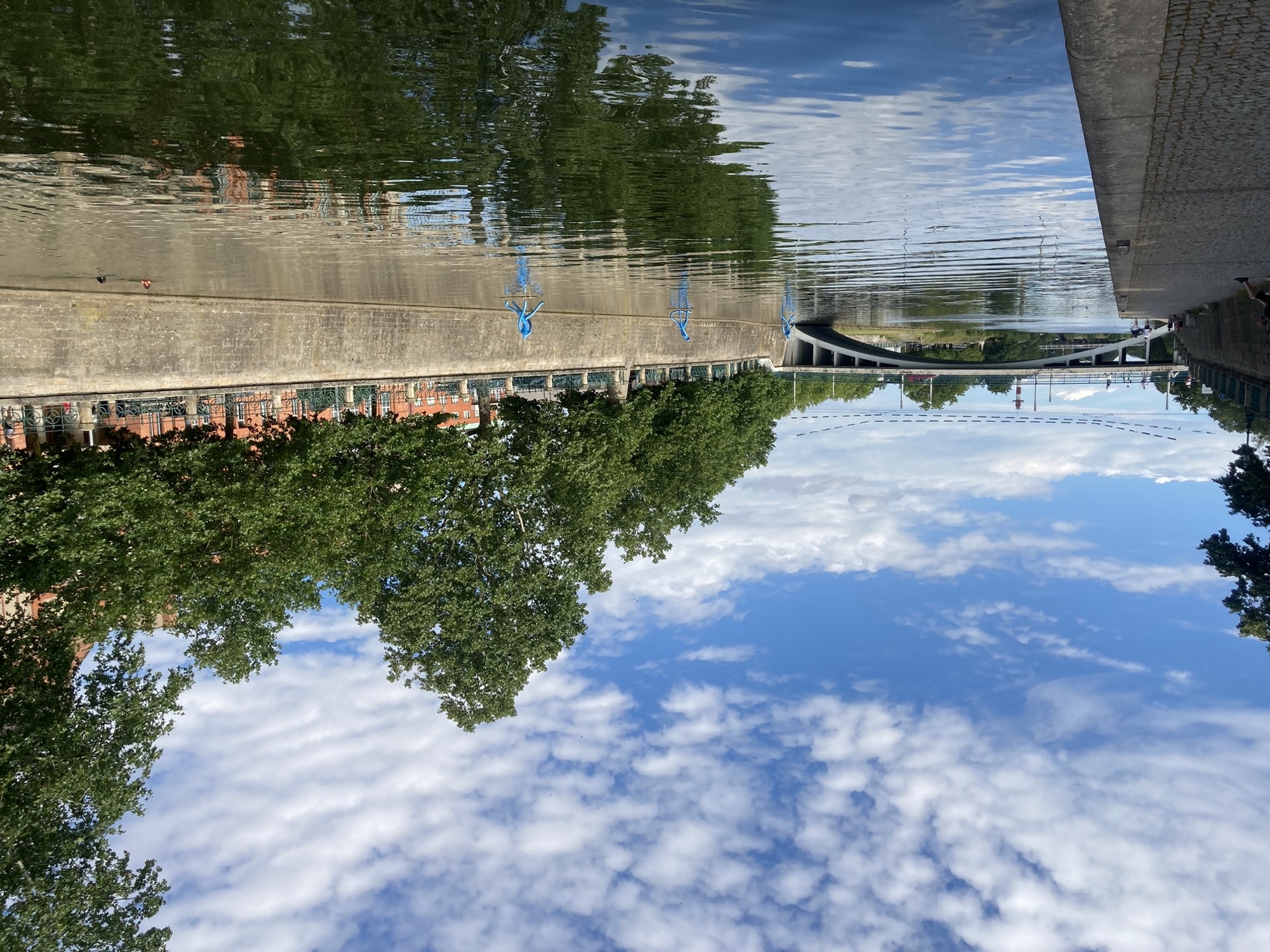 Blick über den Fluss, im Wasser spiegelt sich die Uferpromenade, auf der Wasseroberfläche scheinen drei blaue Statuen zu schwimmen